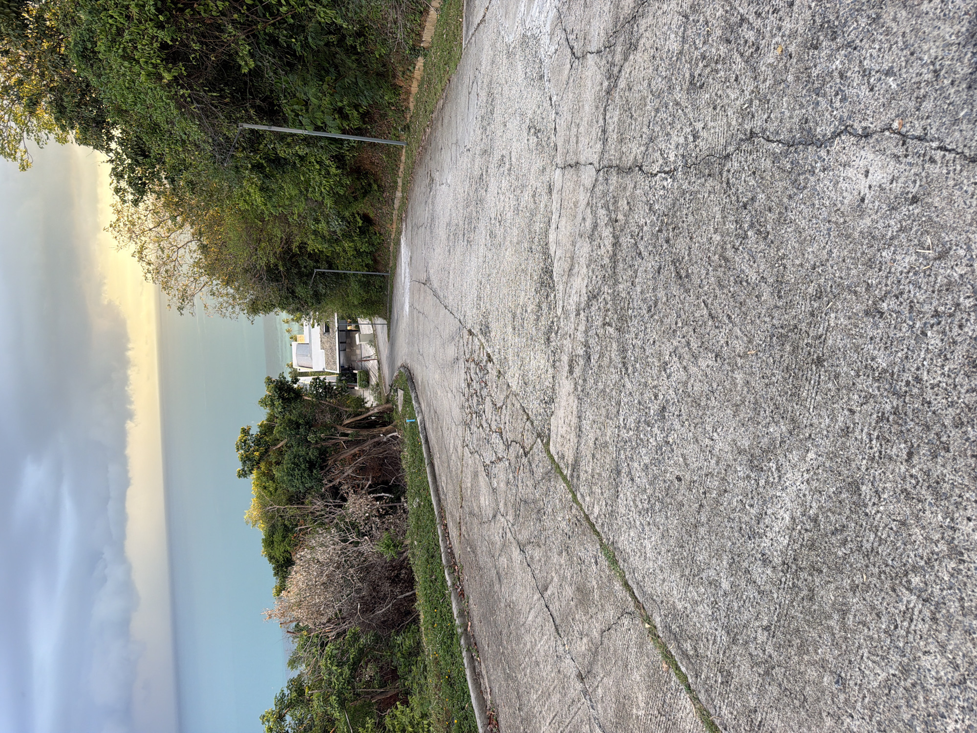 The image shows a cracked concrete road descending a steep hill. Lush green vegetation and trees line the right side of the road, while the left side offers a view of the turquoise ocean under a cloudy sky. In the distance, glimpses of modern white buildings are visible amidst the trees, suggesting a residential or resort area. Two lampposts are positioned along the right side of the road. The scene appears to be in Baan Thong Po, Thailand, given the tropical foliage and coastal view. There are no people or animals visible in the image.