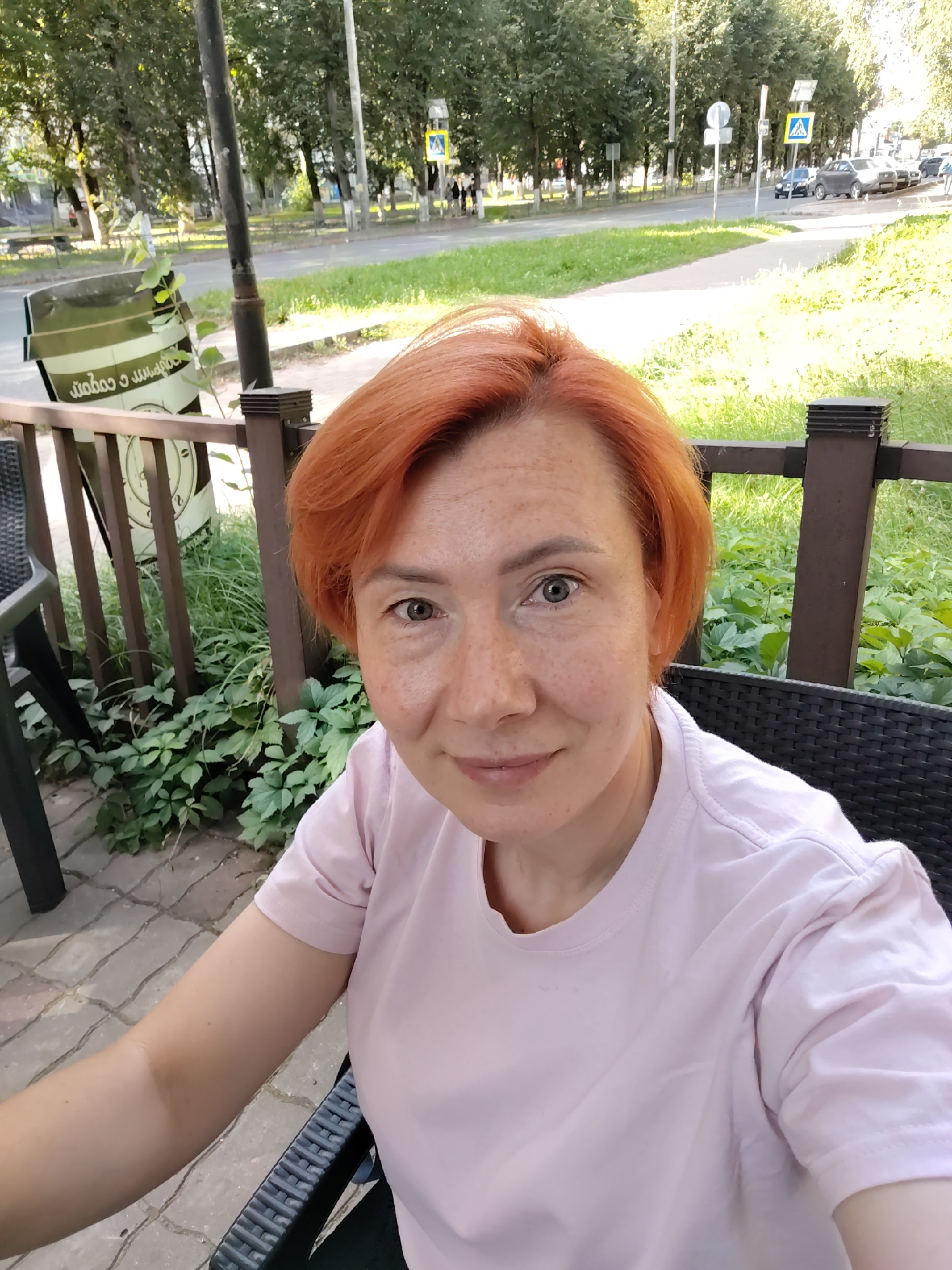 A woman with short, red hair and freckles is taking a selfie in an outdoor cafe. She is wearing a light pink t-shirt and is seated in a black chair. Behind her, there is a wooden railing and lush green plants. In the background, a street with cars and trees is visible, suggesting an urban setting. The overall impression is that of a casual, daytime scene in Konakovo, Russia.