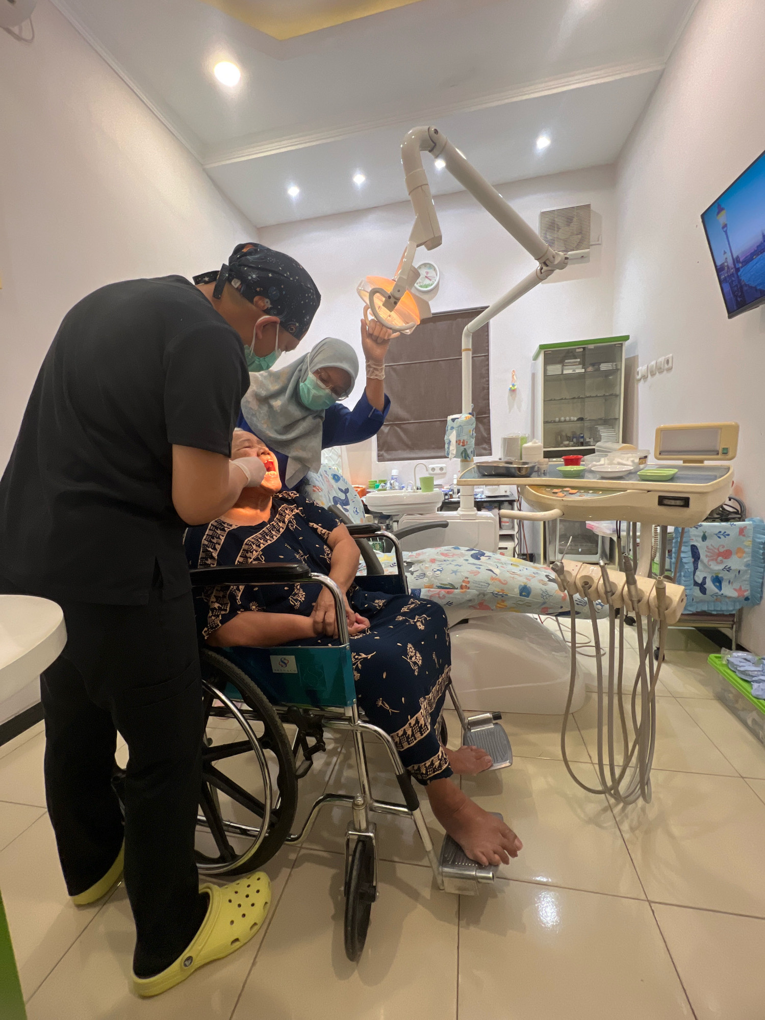 In a brightly lit dental clinic, two medical professionals attend to an elderly woman seated in a wheelchair. The woman, wearing a dark blue patterned dress, has her mouth open as a dentist, dressed in black scrubs and a blue surgical mask, examines her teeth. Another medical professional, a woman wearing a light blue hijab and a green surgical mask, holds a dental light over the patient's mouth, assisting in the examination.
The clinic is equipped with standard dental instruments and furniture. A dental chair is visible, and a tray with various tools rests on a mobile stand beside the patient. Medical cabinets and a sink are also present in the room, suggesting a typical dental examination or treatment setting.
The flooring is tiled with a beige tone, and the walls are painted white, reflecting the artificial lighting. A wall-mounted television displays a picture of a cityscape, possibly an artistic rendition of a European city with a prominent tower. The overall atmosphere suggests a professional and clean environment for dental care. The image does not provide explicit clues to pinpoint the exact location within Magelang, Indonesia, but the scene is consistent with a dental practice anywhere in the city.