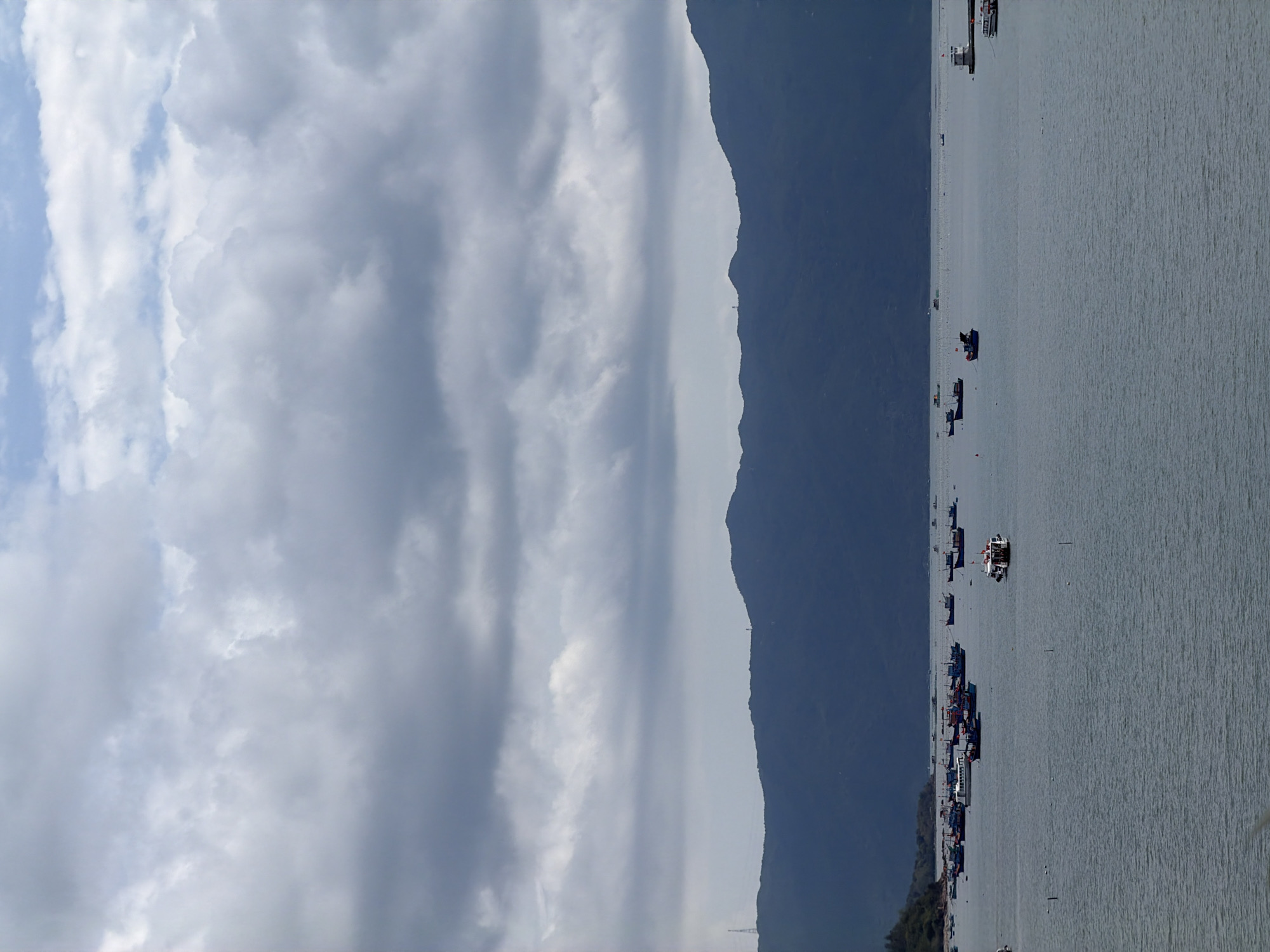 The image depicts a coastal scene in Bac Nha Trang Ward, Vietnam. The sky is filled with dramatic cloud formations, ranging from light to dark gray. A dark, steep mountain range runs vertically down the middle of the image, separating the sky from the sea. On the right side, the water surface is rippled and dotted with numerous boats, ranging in size and color. The boats appear to be anchored or slowly moving along the coast. The coastline is not clearly visible but appears to be populated with structures.