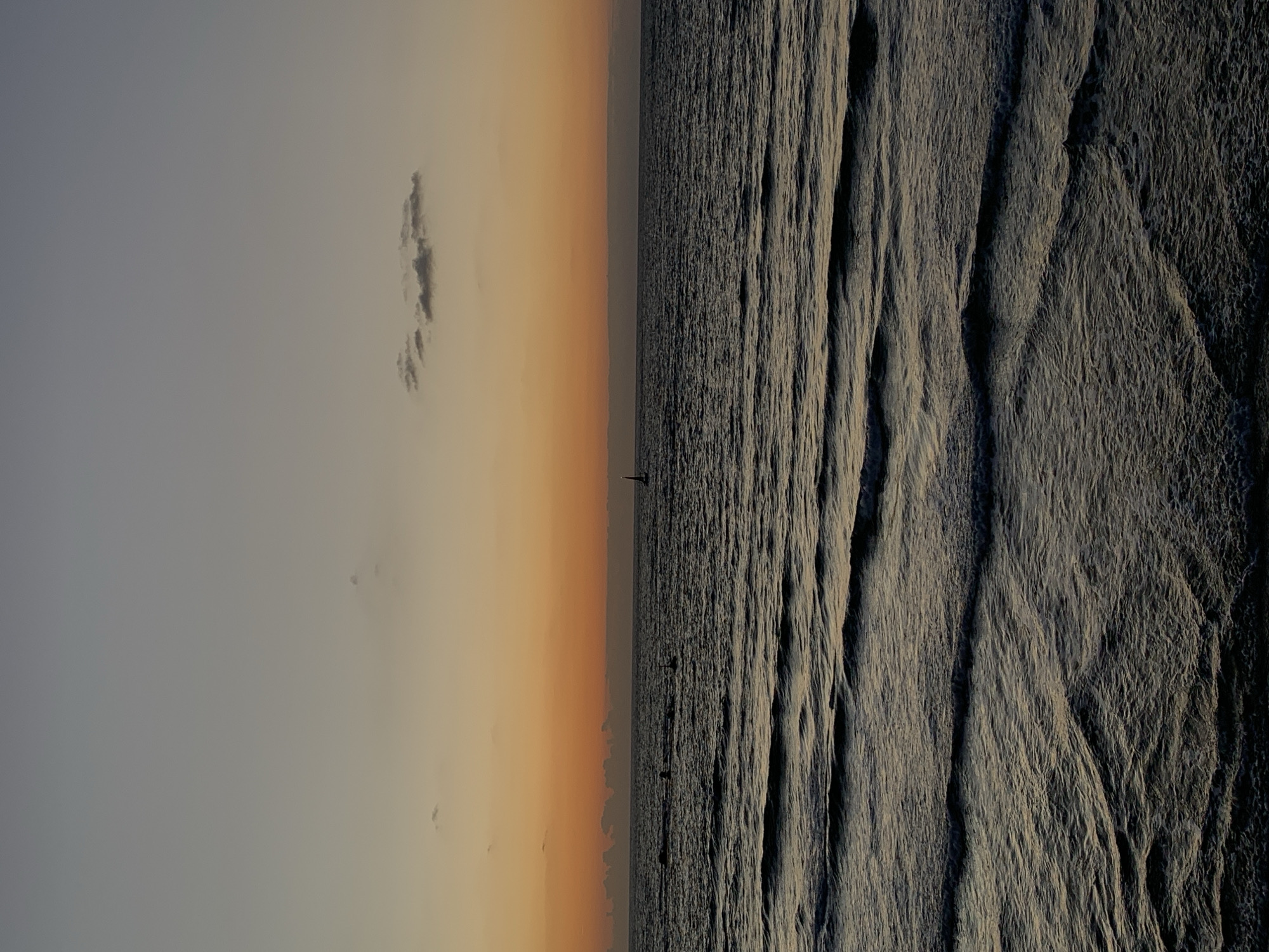The image depicts a seascape at dusk or dawn, likely in Tel Aviv, Israel, although no specific landmarks are visible to confirm this. The left half of the image showcases the sky, transitioning from a light grey to a vibrant orange gradient near the horizon. A few small clouds dot the sky. On the right half, the sea exhibits a textured surface with distinct wave patterns. A small sailboat is silhouetted on the horizon.