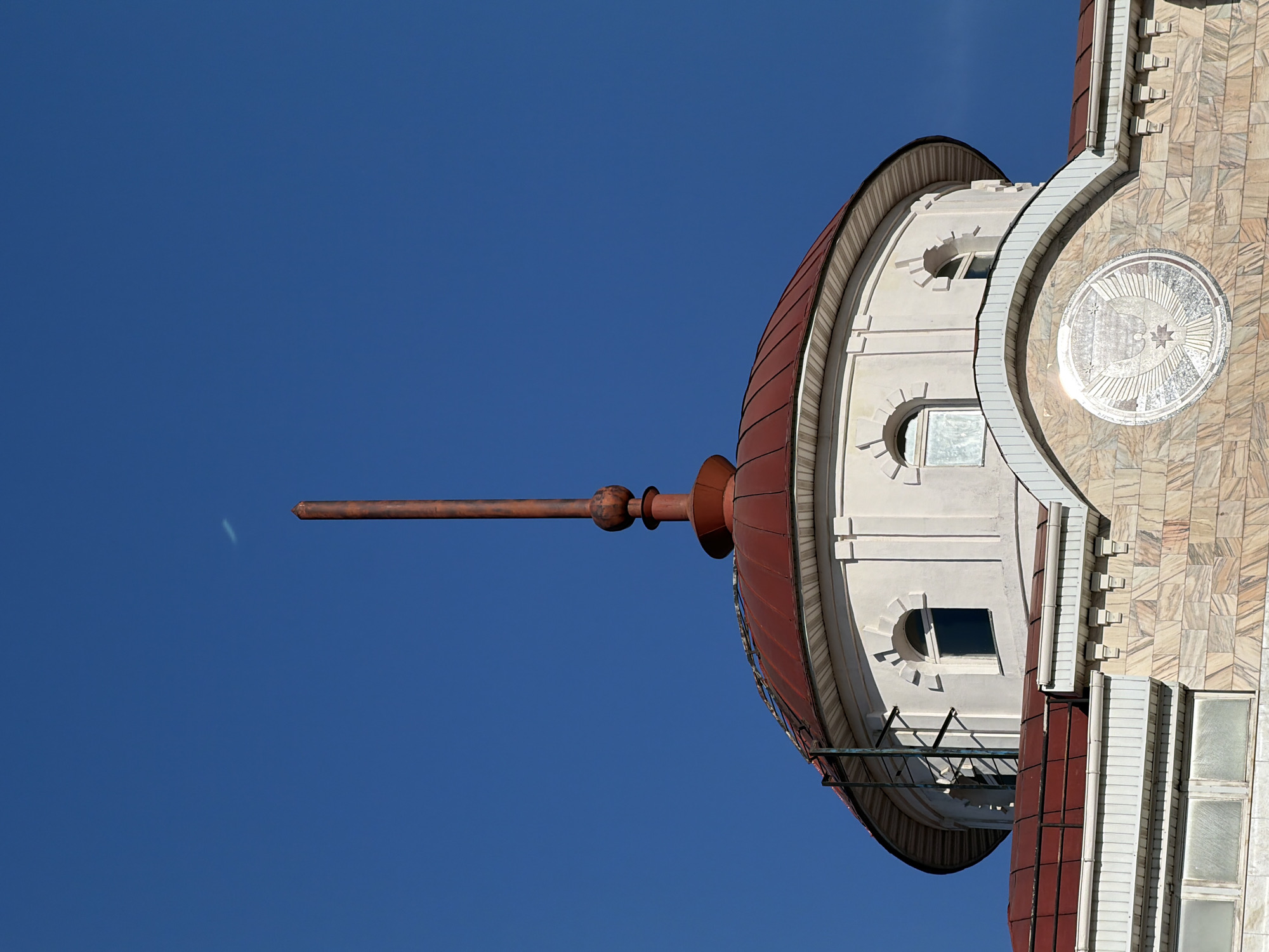 The image shows the dome of the Republican National Library in Izhevsk, Russia, against a clear blue sky. The dome is topped with a long, pointed, rusted-metal rod extending outwards. The dome's structure features rounded windows and decorative architectural elements, including a circular design with a star and crescent moon motif. The building's exterior is a mix of white and textured stone or tile. A portion of the building's facade is visible to the right, with visible windows.