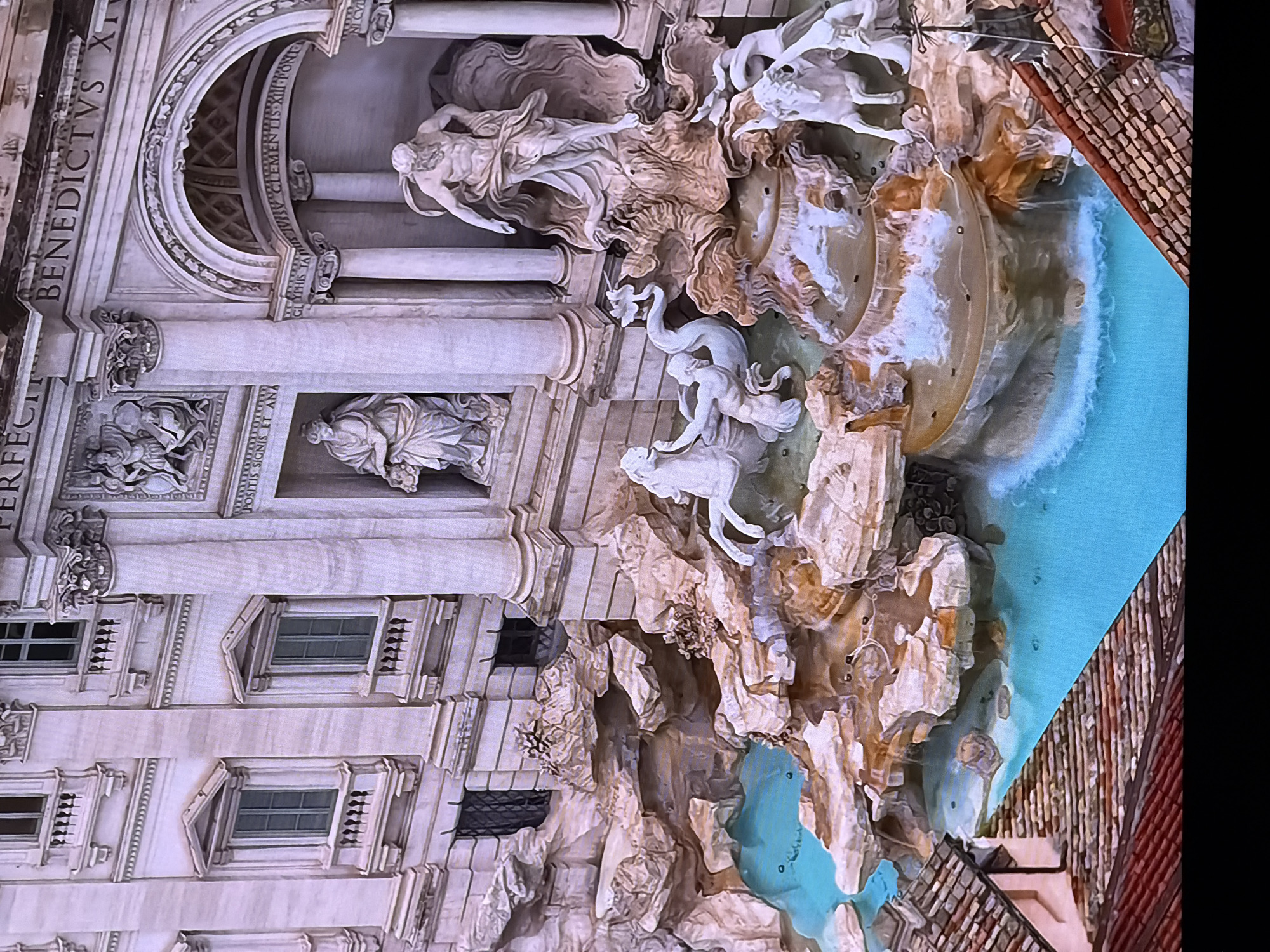 The image shows a high-angle view of the Trevi Fountain in Rome, Italy. The fountain is a large Baroque-style fountain that features a central statue of Oceanus, flanked by two tritons and two winged horses. The water cascades down over rock formations into a large basin. In the background, the facade of the Palazzo Poli is visible, with its numerous windows and statues. The image also shows some red-tiled roofs in the foreground, suggesting a viewpoint from an adjacent building. The lighting suggests it is daytime. There are no people clearly visible in the image, only the sculpted figures of the fountain. The location of Chirchik, Uzbekistan is not represented in this image.