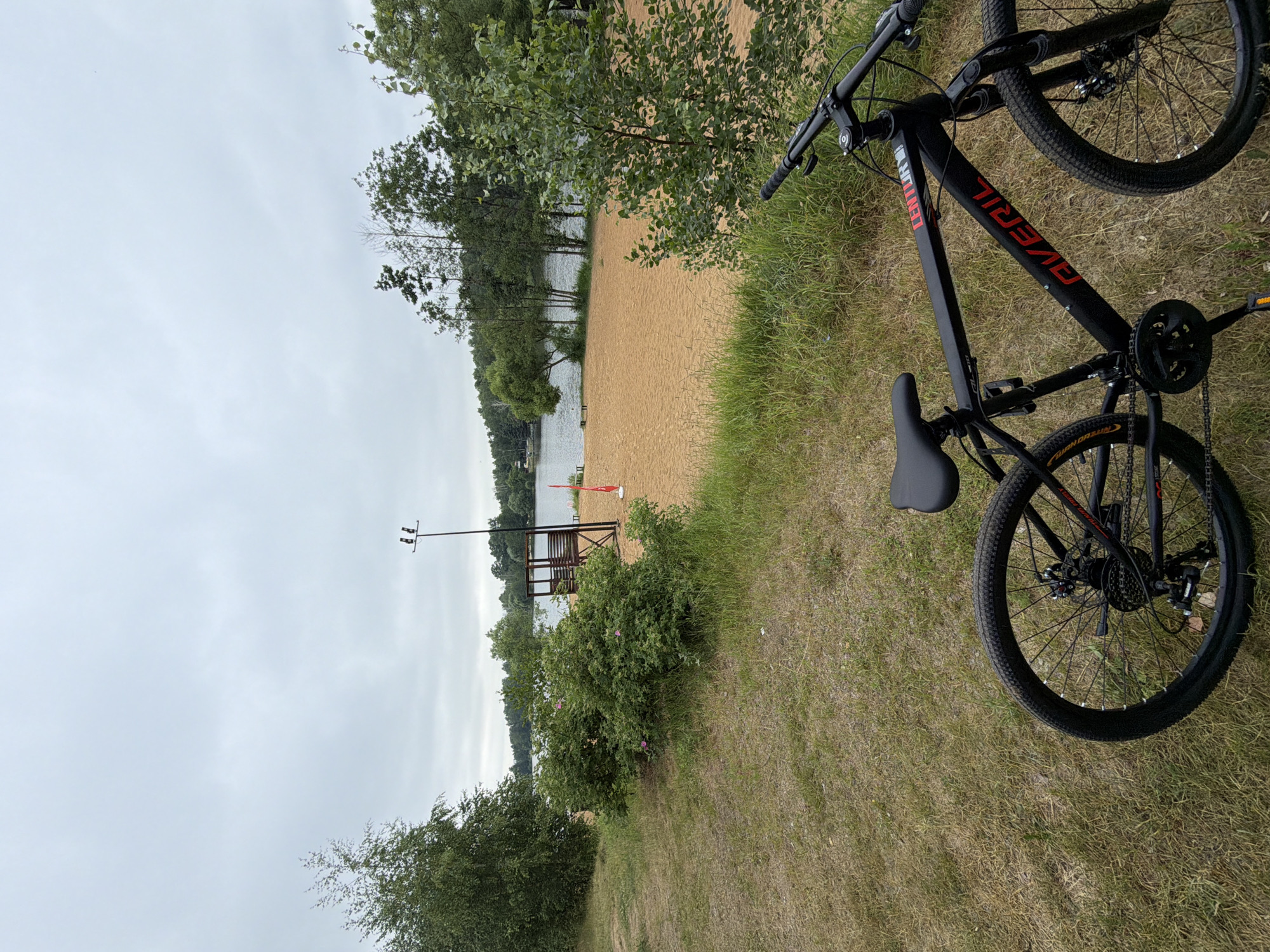 An outdoor, eye-level, full shot shows a black Averil brand mountain bicycle parked in front of a sandy beach on a cloudy day. The bicycle is positioned diagonally in the lower right corner of the frame, facing away from the viewer. Its handlebars are turned slightly to the left. Behind the bicycle, a sandy beach stretches towards a calm lake. A wooden lifeguard stand with a red flag is visible on the beach. Trees line the far side of the lake and dot the landscape. The foreground consists of dry grass and some green vegetation. The sky is overcast, casting a muted light over the scene. The location is identified as Malakhovka, Russia.