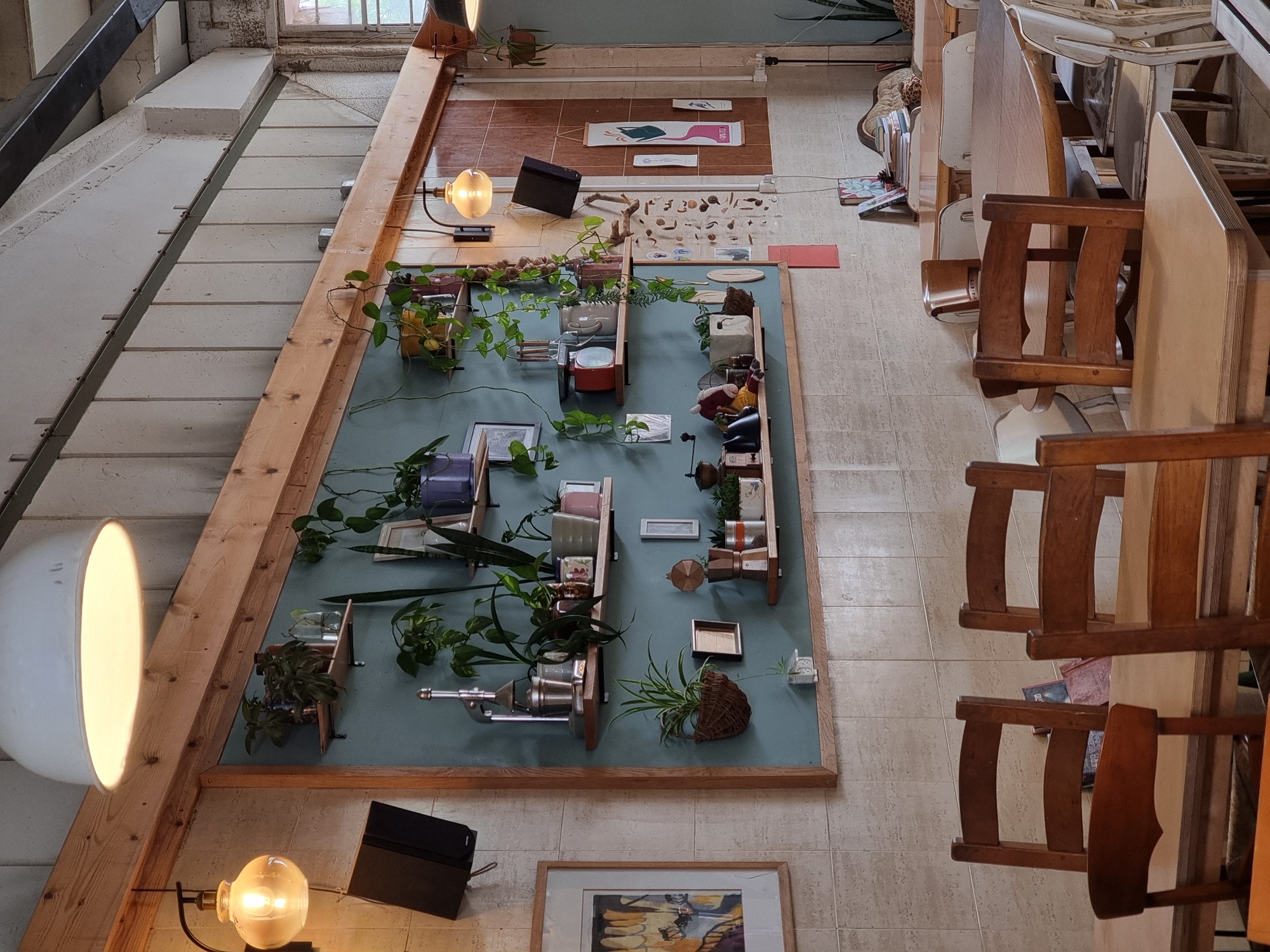 The image shows an interior space in Mitzpe Ramon, Israel. The viewpoint is directed upwards, emphasizing the vertical layout of the scene. A large wall-mounted display dominates the left side of the frame, showcasing an assortment of potted plants, small objects, and framed pictures arranged on a light blue surface with a wooden frame. The display appears to be an artistic installation or a decorative arrangement. Two lamps, one a large dome-shaped light and the other a smaller bulb lamp, illuminate the display. To the right, a series of wooden chairs and tables are stacked and arranged, suggesting a storage area or a workspace. The floor is tiled, and glimpses of an exterior window and architectural details are visible at the top of the frame. The overall atmosphere is that of a studio, workshop, or gallery setting.