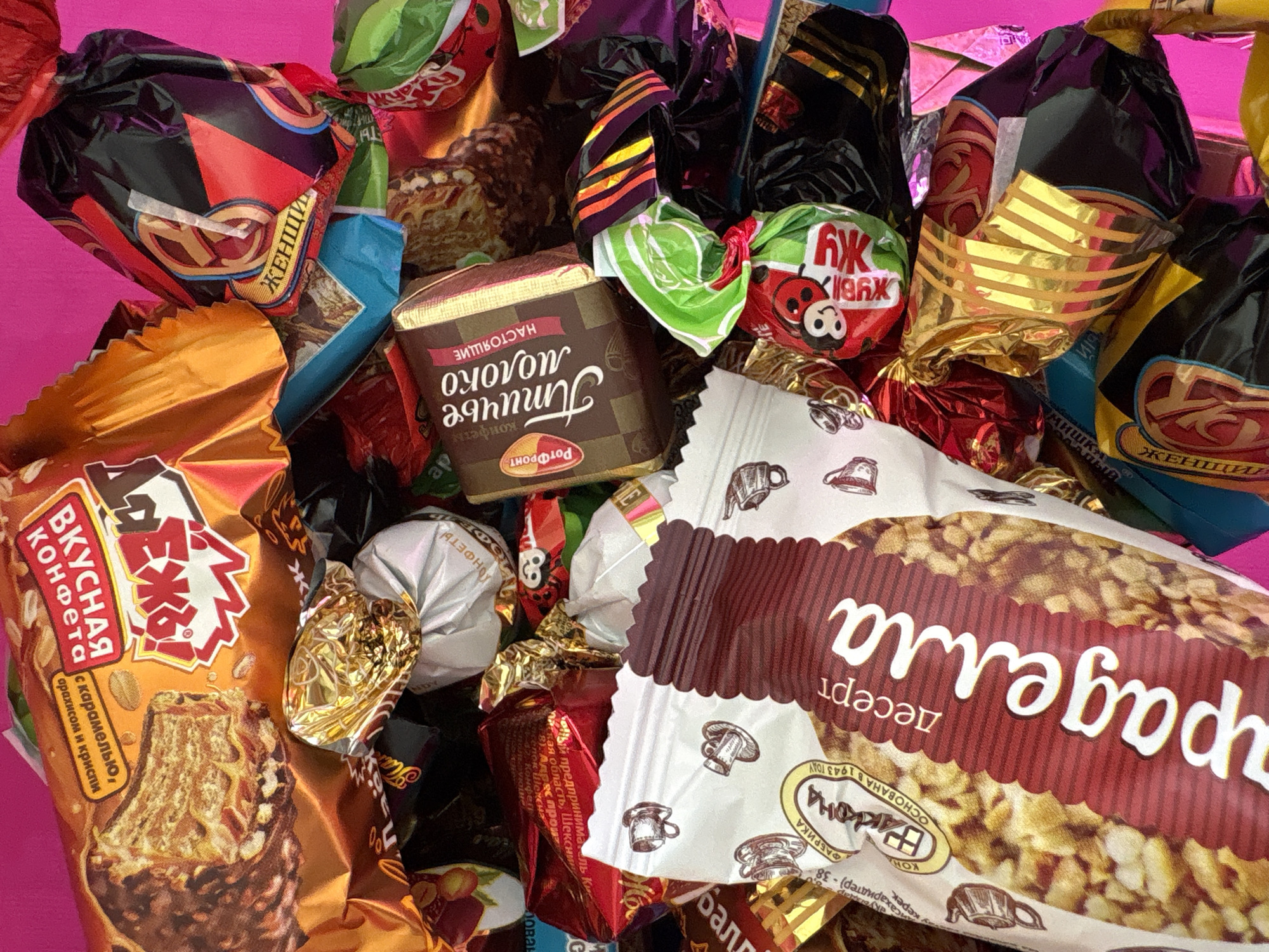 A close-up shot displays an assortment of Russian candies against a bright pink backdrop. The candies vary in shape, size, and packaging design, with visible text in Russian. Prominent brands include "Akko", "Rort Front", "Aparagella" and "Bkusnaya Konfeta". The "Мишки" brand is partially visible. The image includes details like product names ("НАСТОЯЩИЕ молоко") and phrases such as "ВКУСНАЯ конфета" (delicious candy) along with information about the manufacturer and ingredients. One candy packaging mentions the confectionary factory "ФАБРИКА ВаКкоНа" founded in 1943. The text also mentions the businessman Kis of Sheksna, Vologda region. No people or actions are visible. The environment is limited to the close proximity of the candies and the pink backdrop. The location in Izhevsk, Russia, is not discernible from the image.