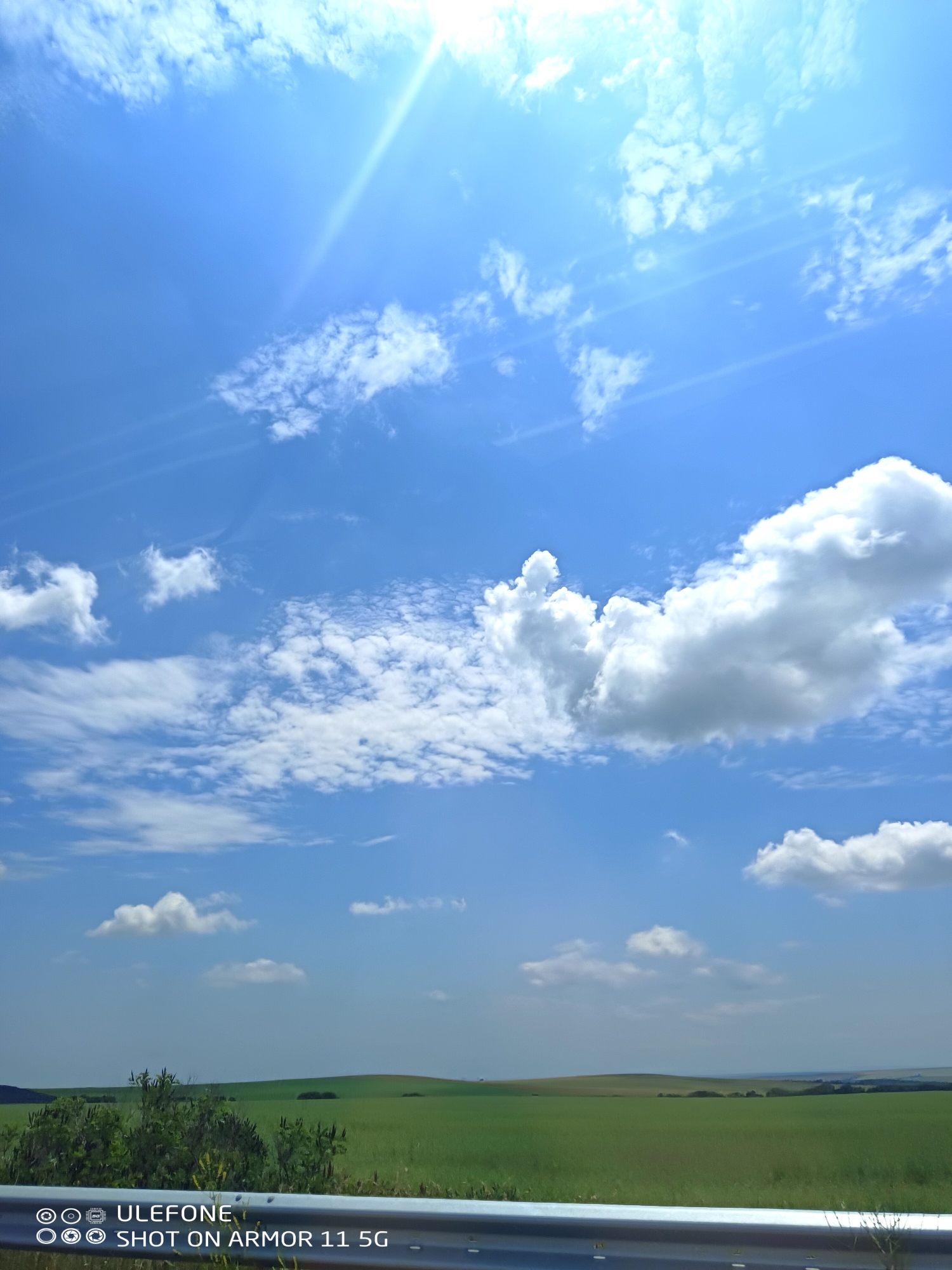 The image displays an expansive outdoor landscape under a bright, clear blue sky with scattered white clouds, characteristic of Kozare, Bulgaria. The sun, positioned in the upper right quadrant, emits a visible lens flare and prominent sun rays. The sky transitions from a lighter blue near the horizon to a deeper blue overhead, hosting various cloud formations including fluffy cumulus clouds and more diffuse, wispy clouds. The lower portion of the image reveals rolling green hills and fields, indicating an agricultural or rural setting. Different shades of green suggest varied vegetation or crops across the gently undulating terrain. In the immediate foreground, a silver metallic guardrail, consistent with a roadside barrier, spans the width of the image, partially obscuring some dark green shrubs and sparse vegetation directly in front of it. No individuals, animals, or specific activities are present. A digital watermark in the lower left corner reads "ULEFONE SHOT ON ARMOR 11 5G".