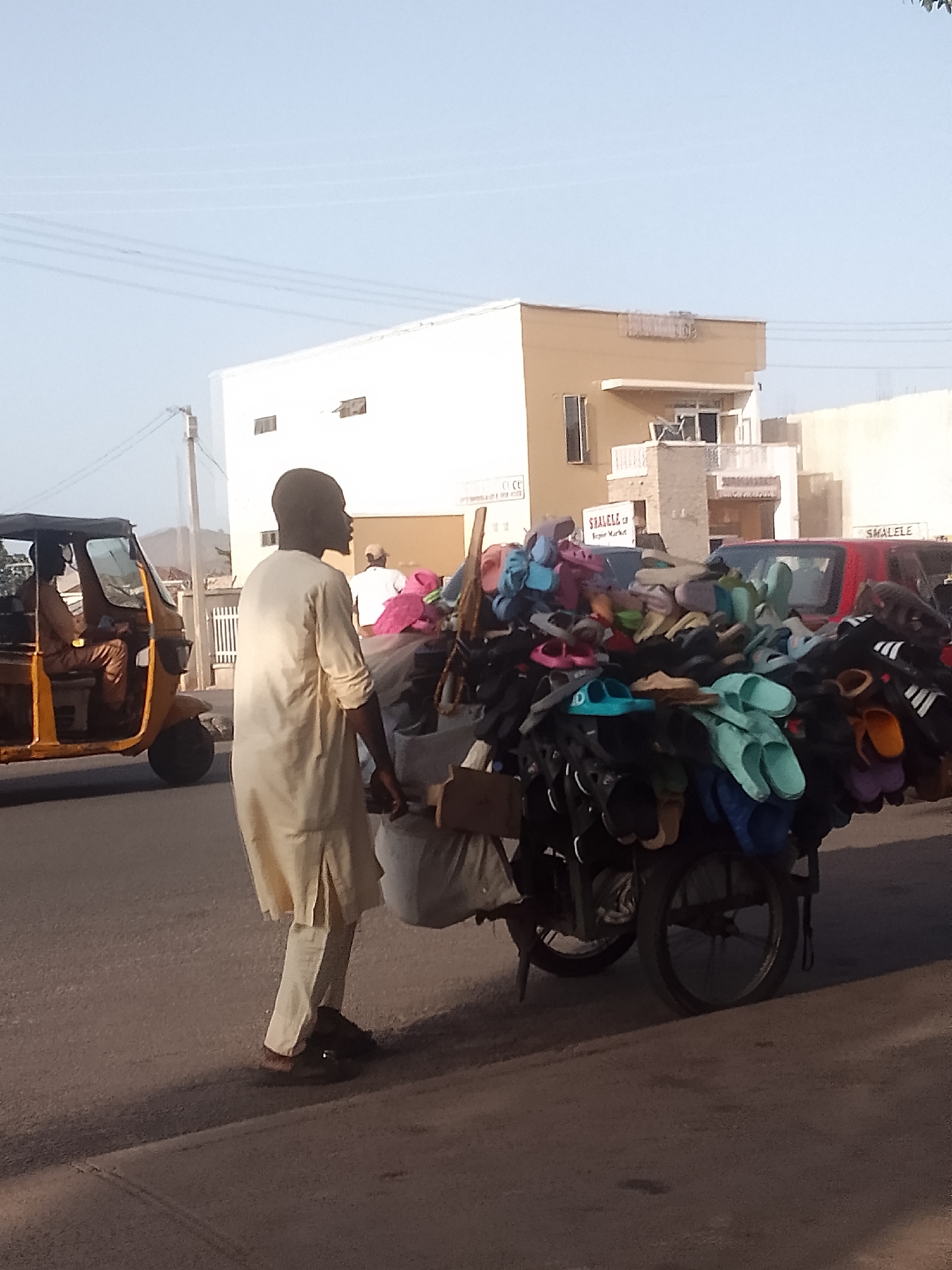 A man wearing a long, light-colored tunic and dark shoes is walking on a paved road in Pantame, Nigeria. He is pulling a two-wheeled cart heavily loaded with an assortment of colorful sandals and flip-flops. To his left, a yellow three-wheeled auto-rickshaw is parked, with a person visible inside. In the background, there's a white building with a smaller tan-colored structure on top, bearing the word "SMALELE". A red vehicle is also visible behind the cart. The overall scene appears to be a street in a residential or commercial area during daylight.