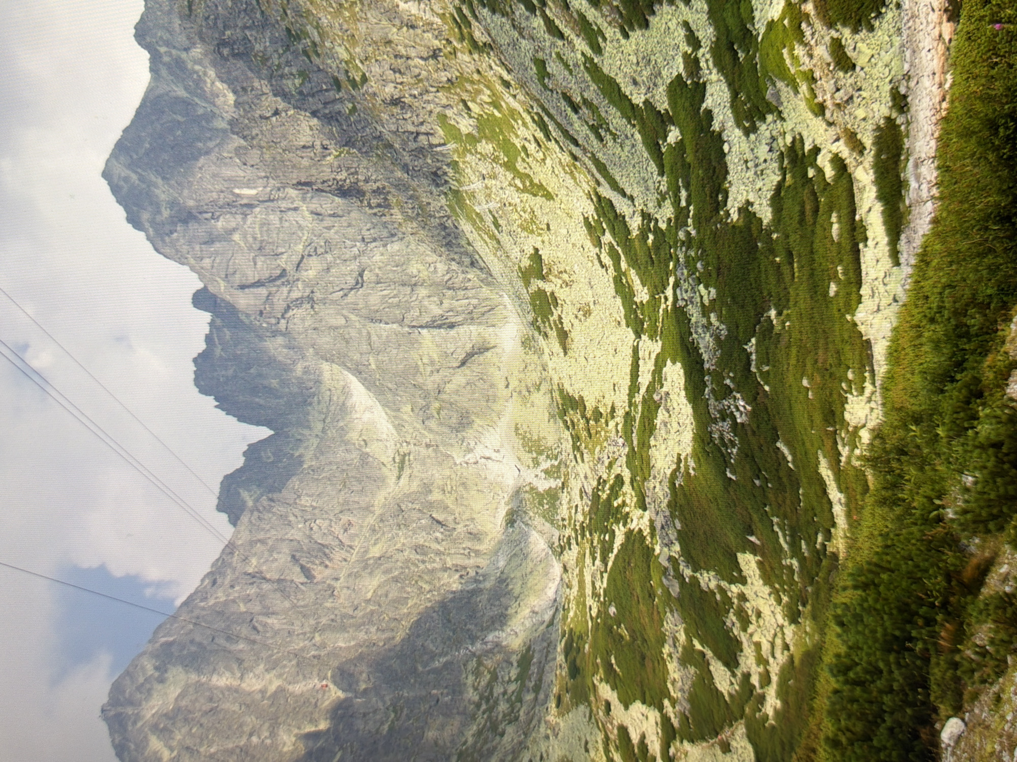 The image shows a mountainous landscape with steep, rocky slopes and patches of green vegetation. Two cables traverse the sky, suggesting a cable car or ski lift system. The terrain appears rugged and natural, with sparse trees in the foreground and large rock formations dominating the background. The lighting indicates it is daytime, with a mix of sunlight and clouds. The scene does not depict any urban elements or features that would suggest a location within the city of Kyiv, Ukraine.