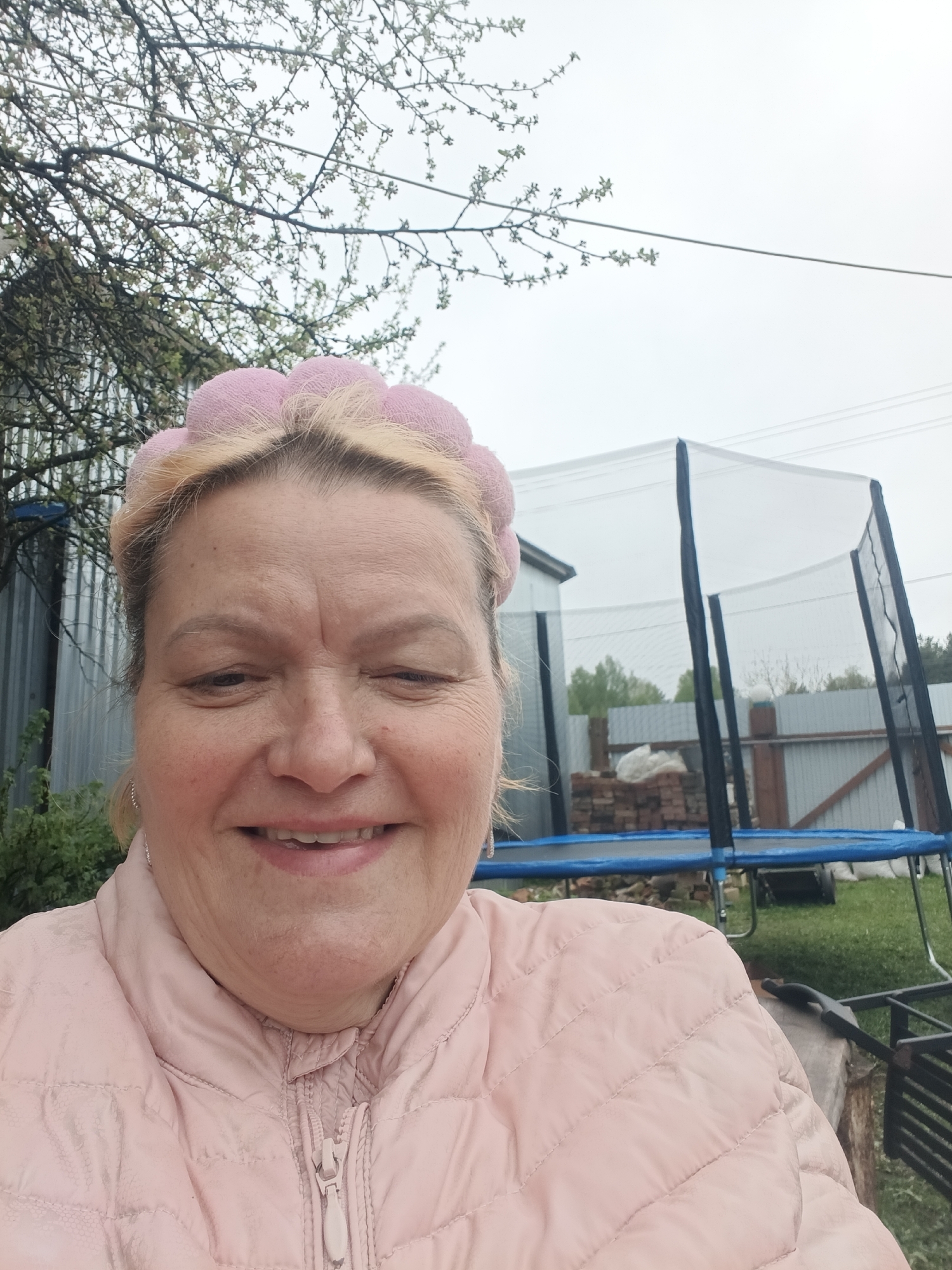 A woman with a pink headband and jacket is smiling at the camera. In the background, there's a trampoline, a metal fence, a building, and trees. The photograph appears to be taken outdoors, possibly in a backyard. The location is Красный Маяк, Russia.
