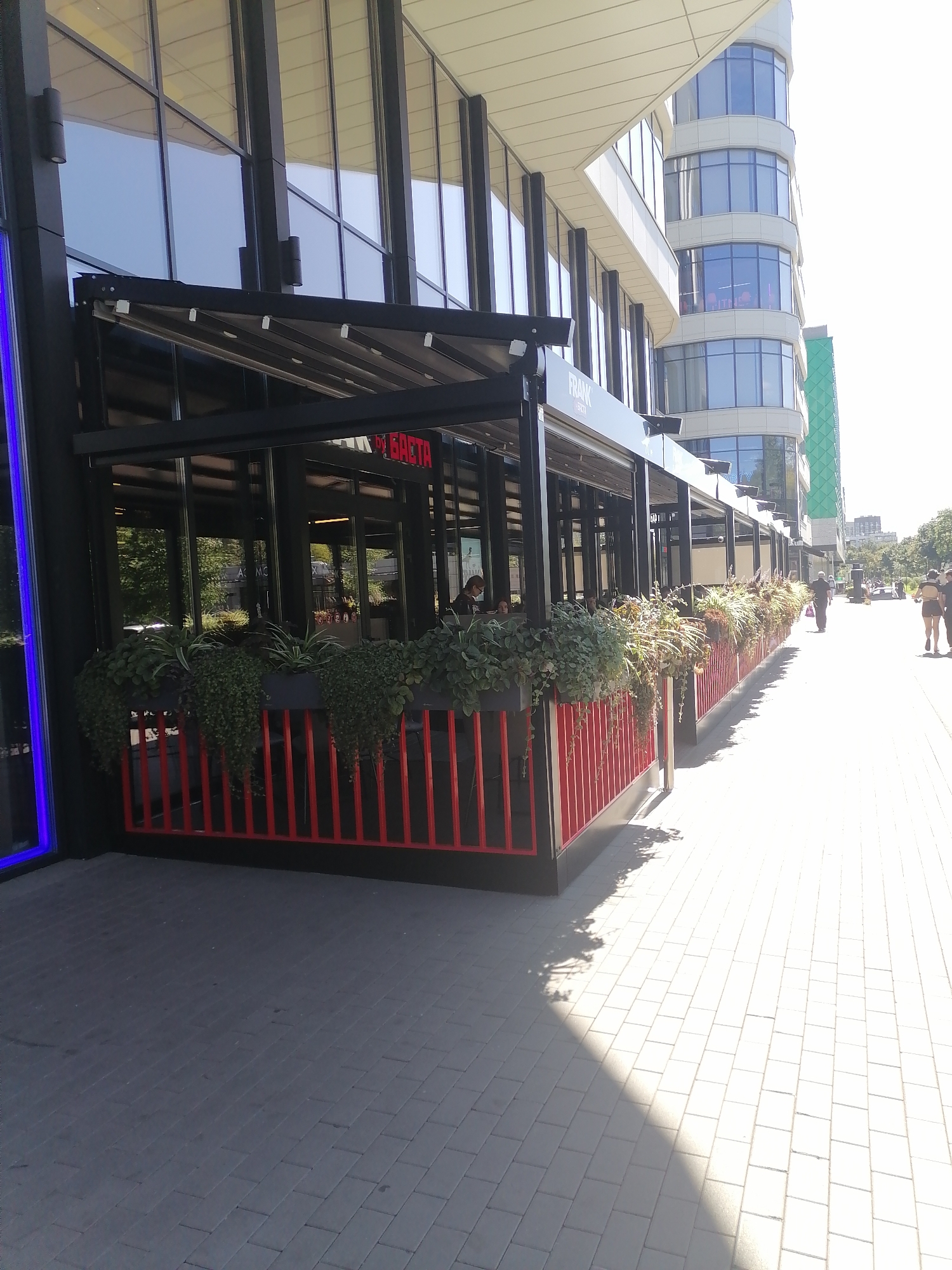 The image shows a cafe with an outdoor seating area on a sunny day. The cafe has a black awning with a retractable roof, and a red metal fence with planters filled with greenery in front of it. The cafe has large glass windows and doors. In the background, there is a modern, multi-story building with a curved facade and numerous windows. A sidewalk paved with light-colored rectangular stones extends from the cafe. Several people are visible walking on the sidewalk in the distance. The scene appears to be located in a city, likely Moscow, Russia, given the architectural style and general urban setting.
