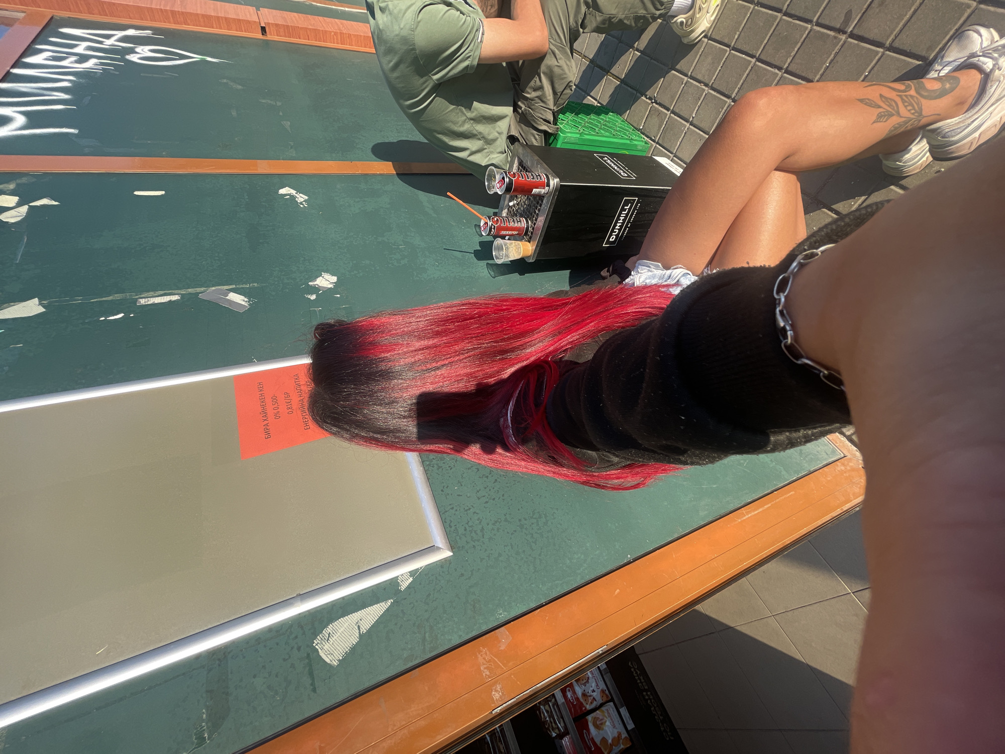 The image depicts a street scene in Sofia, Bulgaria, likely during daylight hours with strong sunlight casting shadows. In the foreground, a person with vibrant red and black hair is seated. They are wearing a dark top and light-colored shorts, and a silver chain bracelet is visible on their wrist. A tattoo of a plant with a serpent is visible on their lower leg.
To the right of the seated person, a black Dunhill branded trash receptacle is present, with two open cans of drink and a straw on top, suggesting a break or refreshment. A person in a green t-shirt and olive green pants is seated behind the trash receptacle. Green plastic crates are stacked to the right of the receptacle.
The background features a dark green wall with white graffiti, including the name "МИЛЕНА" and a heart symbol. A bright orange-yellow frame or structure runs vertically to the left of the green wall. A sign with Cyrillic text in orange is affixed to a lighter-colored surface behind the person's head. A display shelf with various products, possibly snacks, is visible to the far left, partially obscured by the orange-yellow structure. The ground is paved with square tiles.
