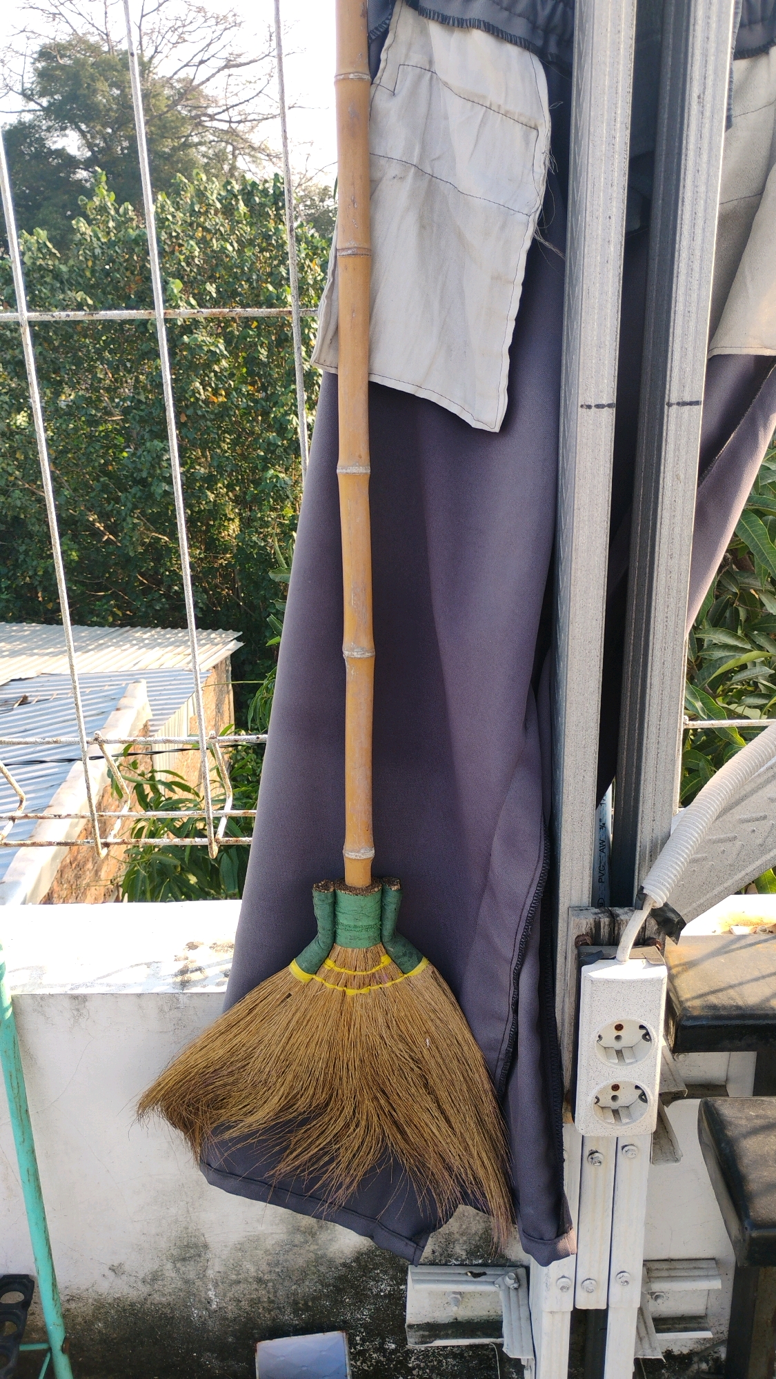 In Gumpang, Indonesia, a close-up shot captures a traditional broom with a bamboo handle and natural bristles. The broom is leaning against a dark gray fabric, possibly part of a garment or covering. Above the broom, a light-colored cloth with stitched lines is attached to the bamboo handle, and further up, a section of a metal grate or fence is visible. In the background, lush green foliage and a portion of a metallic roof structure are present, suggesting an outdoor setting, potentially a rooftop or balcony. The scene is illuminated by natural light, casting soft shadows.
