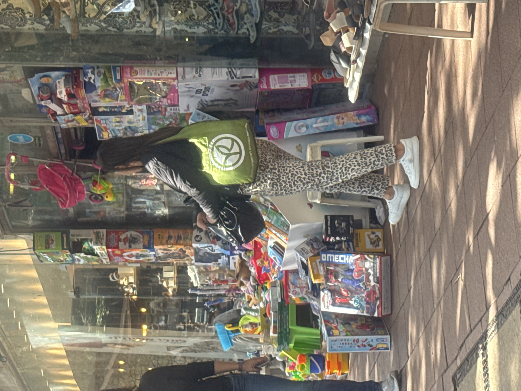 A medium-shot photograph, taken in the city of Ramat Gan, Israel, depicts a street scene reflected in a shop window. The reflection captures a person walking on a sidewalk with a green shopping bag. They are wearing leopard-print pants and white sneakers. The shop window displays various toys, including toy cars, dolls, and building blocks. The reflection also shows sections of the street, including portions of the sidewalk, and other pedestrians.