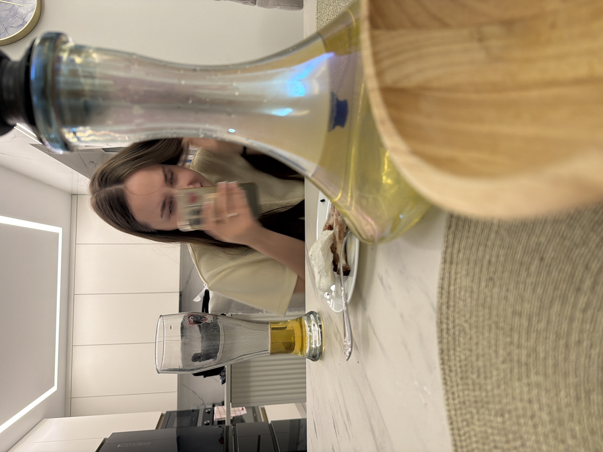 A woman is taking a photograph with her mobile phone of a glass of beer, a plate of food, and a large glass container on a table in a modern kitchen in Молодёжный, Russia. The kitchen has white cabinets and stainless steel appliances. There is a clock on the wall.