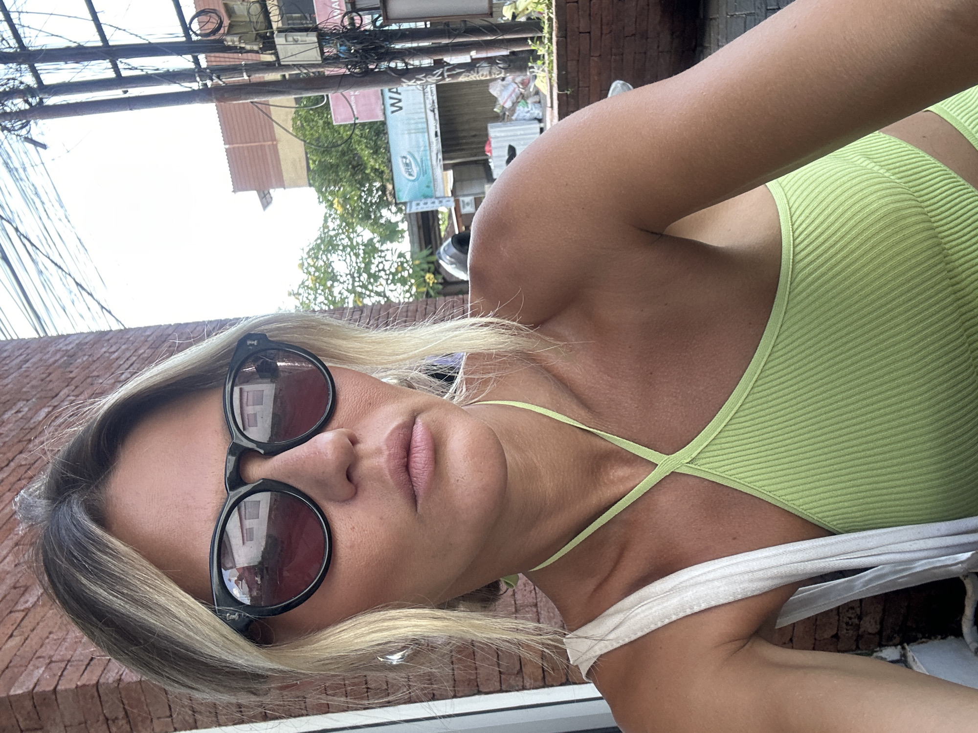 A young, fair-skinned woman with blonde hair pulled back is taking a selfie outdoors. She is wearing large, black-rimmed sunglasses with red lenses, a lime green ribbed tank top with a halter neck, and a white canvas tote bag over her left shoulder. She is outdoors with a brick wall on the left and a street scene in the background with power lines, utility poles, trees, and buildings. The location is identified as North Kuta, Indonesia. The lighting suggests it is daytime with bright sunlight.