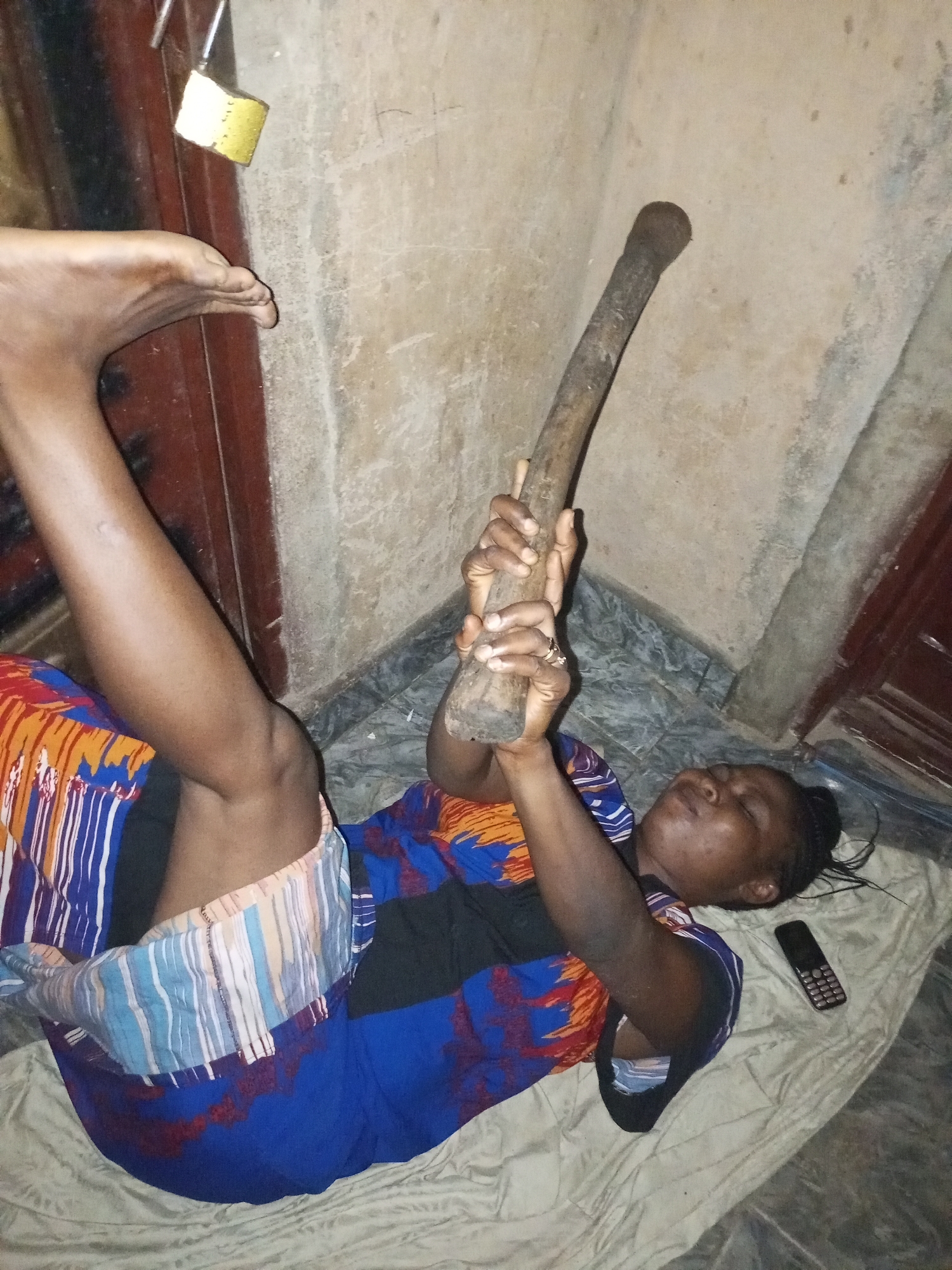 A woman is lying on her back on a beige mat, with her legs extended upwards. She is wearing a blue, orange, and white patterned dress. In her hands, she is holding a long, dark wooden stick. Her eyes are closed, and her head is tilted back. A black mobile phone is lying on the mat to her right. Behind her is a beige wall with dark brown vertical panels on the left and right sides of the image. A yellow padlock is attached to the left panel. The floor is tiled with dark gray and lighter gray diamond-shaped tiles. The scene appears to be an indoor space in a residential area in Tempure, Nigeria.