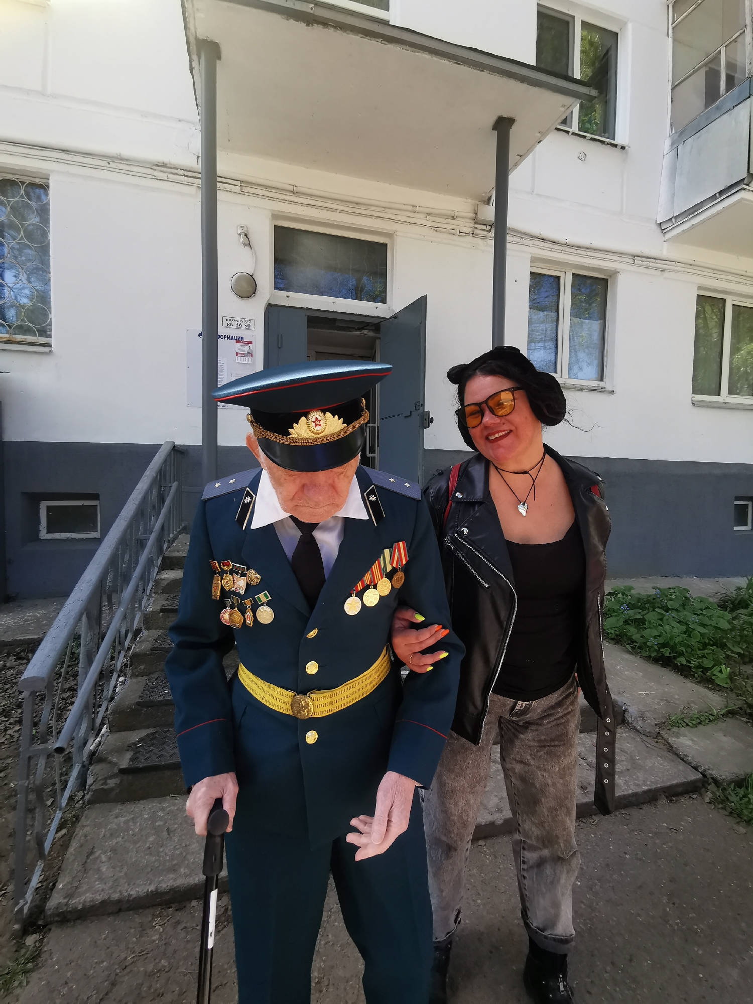 An elderly man in a dark blue military uniform with numerous medals is walking down concrete steps. He is using a black cane and is being assisted by a woman. The woman has a black leather jacket, dark jeans, and orange tinted glasses. She is holding the man's arm. The background shows a light gray building with white trim and windows. A sign in Russian is visible on the building. The location is Chaikovsky, Russia.