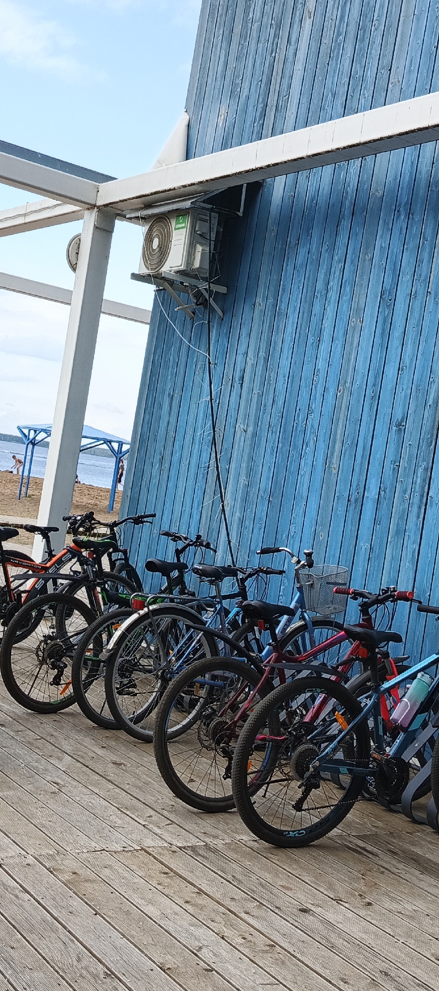 A row of bicycles is parked on a wooden boardwalk in front of a bright blue wooden wall. The bicycles are of various colors, including red, blue, and black, and some have baskets. In the background, the beach and a body of water are visible, with a few people walking near blue beach shelters. An air conditioning unit is mounted high on the blue wall. The sky is blue with some clouds, suggesting it is daytime and the weather is fair. The scene appears to be a recreational area, possibly a beach resort or park. The overall mood is relaxed and summery. No visible text is present on the bicycles or the surroundings.