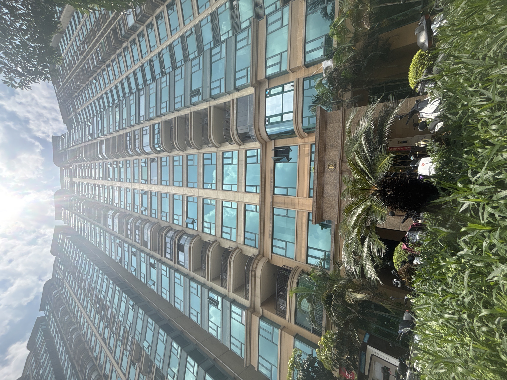 The image shows a low-angle view of a tall residential building in Nanhai District, China, on a sunny day with a partly cloudy sky. The building features numerous floors with glass-paned windows and balconies. In the foreground, lush green vegetation, including tall grass and palm trees, partially obscures the view of the building's base. Several parked motorcycles are visible amongst the plants. The building's facade is a light brown color, and the windows have a blue tint, reflecting the sky. Sunlight streams down from the upper right of the frame, creating lens flare. The overall scene depicts an urban residential environment.