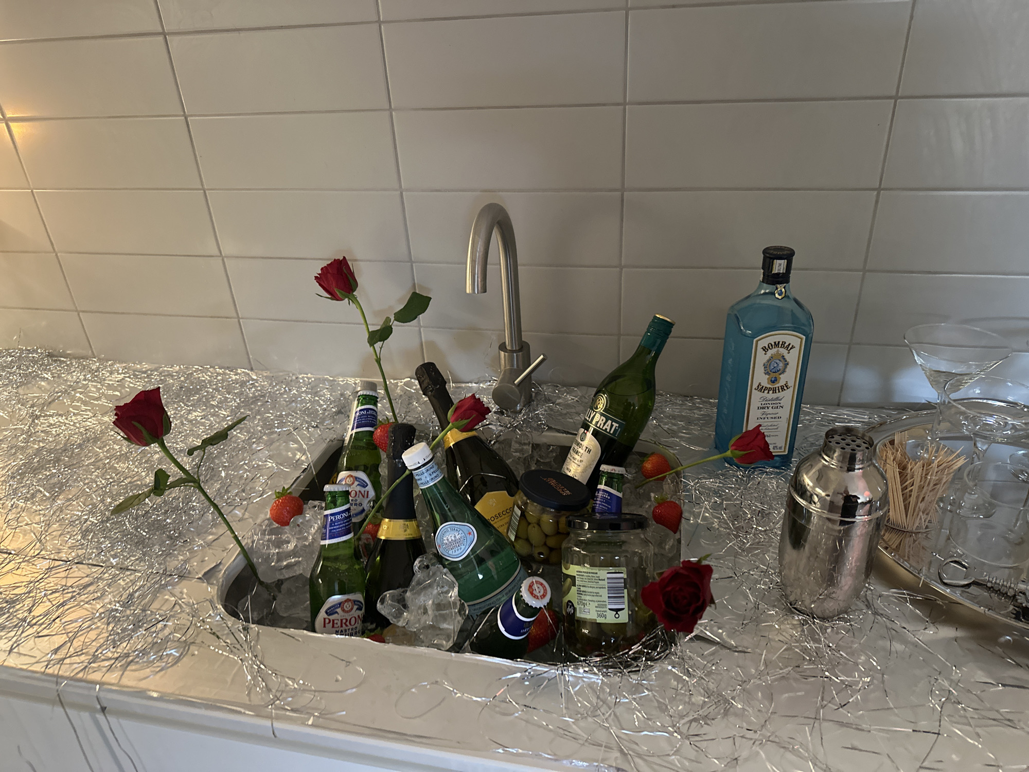 The image displays a festive bar setup, likely for a celebration. The scene features a sink filled with ice, containing various beverages like Peroni beer, Prosecco, Pellegrino sparkling water, and bottles of dry vermouth. Jars of olives and cocktail ingredients are also nestled within the ice. Red roses and strawberries are scattered among the bottles for decoration. A bottle of Bombay Sapphire gin, a cocktail shaker, martini glasses, and toothpicks are situated on a silver tray nearby. The backdrop consists of a light-colored tiled wall, suggesting a modern kitchen or bar area. The overall ambiance suggests a luxurious and celebratory setting. There are no people in the image. The setting appears to be in a domestic location.