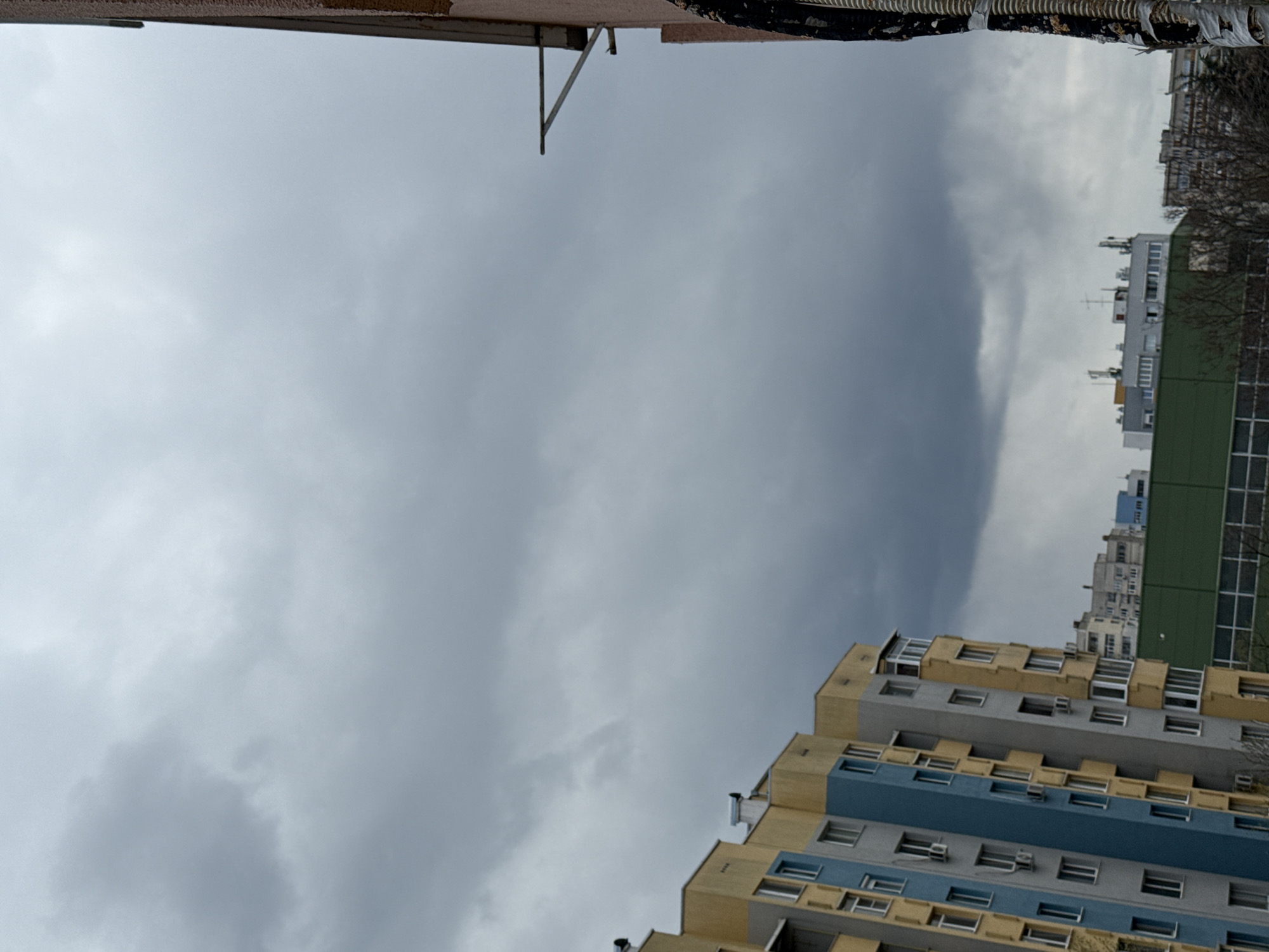 The image is a low-angle shot of a cloudy sky above several buildings. The buildings include a high-rise with blue and yellow detailing, another with a green section, and other structures in the distance. The perspective suggests the photo was taken from a lower vantage point within an urban environment, potentially in Stara Zagora, Bulgaria.