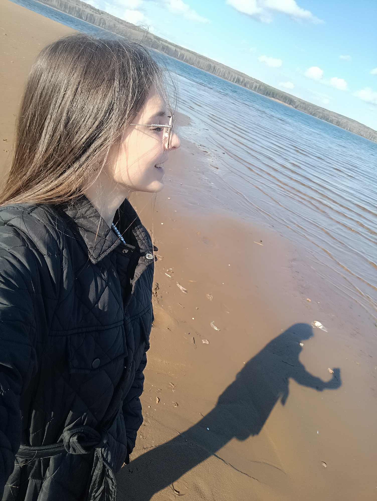 A woman stands on a sandy beach beside a body of water under a bright sky. The location is Unknown, Unknown. The woman is facing to the right of the frame, wearing a black quilted jacket and eyeglasses, her long brown hair falling over her shoulder. Behind her, a wide expanse of water stretches to the horizon, lined with trees in the distance. The sky is blue with scattered clouds. A shadow cast by the woman extends across the sand in front of her.