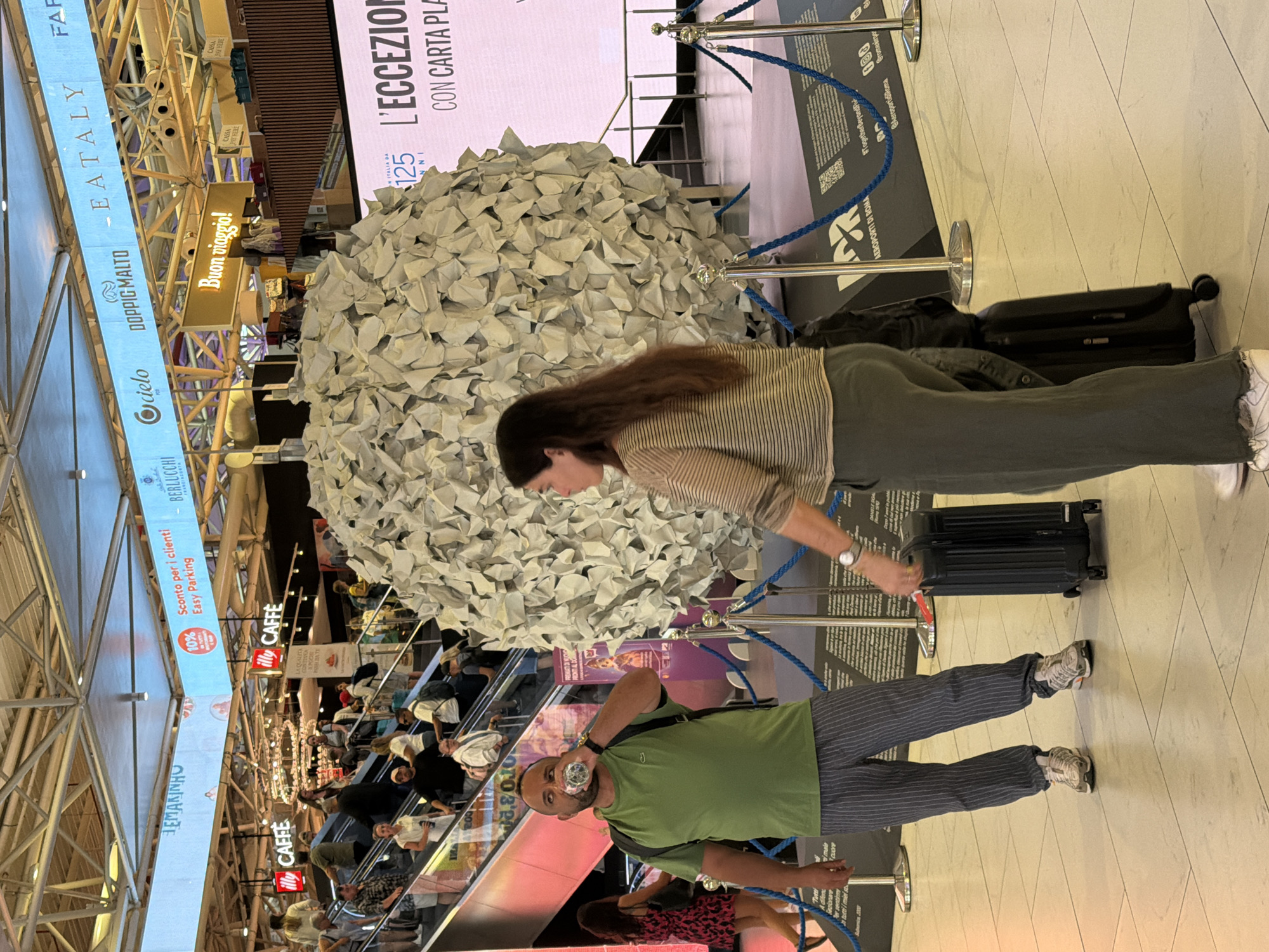 In Fiumicino, Italy, at what appears to be a transportation hub such as an airport or train station, a man in a green t-shirt and striped pants drinks from a water bottle. He stands to the left of a large, spherical sculpture made of crumpled white paper. To his right, a woman with long brown hair, wearing a striped shirt and green pants, stands beside a black rolling suitcase. Behind them, an escalator carries passengers upwards, and various commercial advertisements and signage are visible, including "EATALY," "CAFFE," and "Buon viaggio!" The environment features tiled flooring and overhead structural elements.
