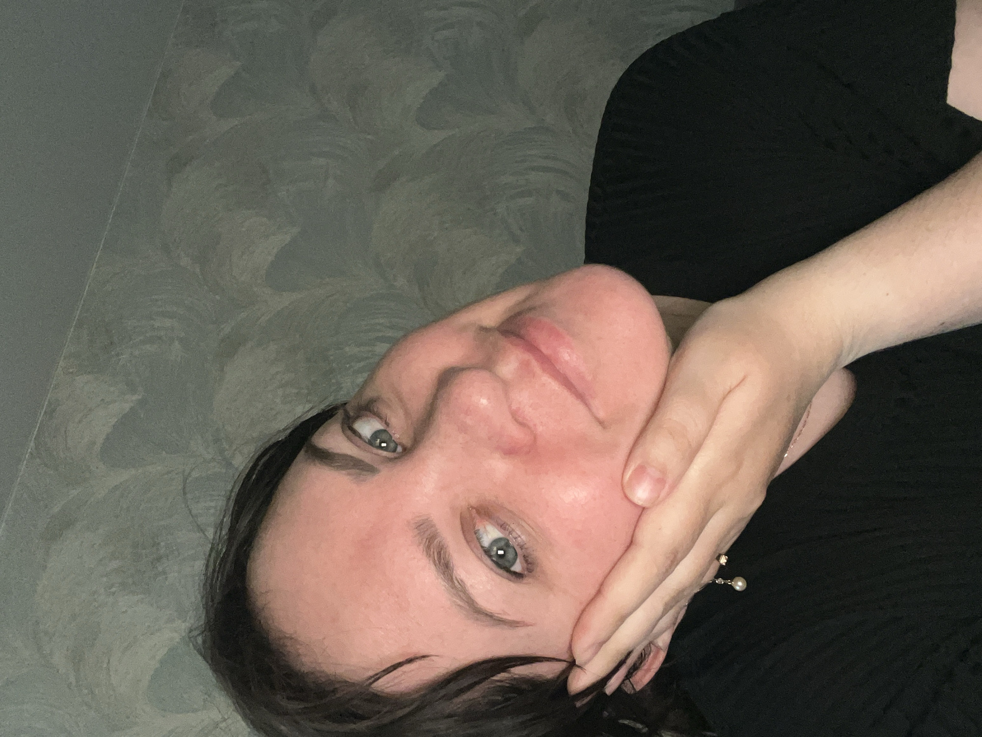 A woman with fair skin, dark hair, and light-colored eyes is shown in a close-up selfie. Her face is angled, with her hand partially covering her cheek and jawline. She is wearing a black top, and a bracelet with a dangling pearl is visible on her wrist. The background appears to be a wall with a textured, wavy pattern in shades of gray. The location is not discernible as Noviy, Russia based on the provided visual information.
