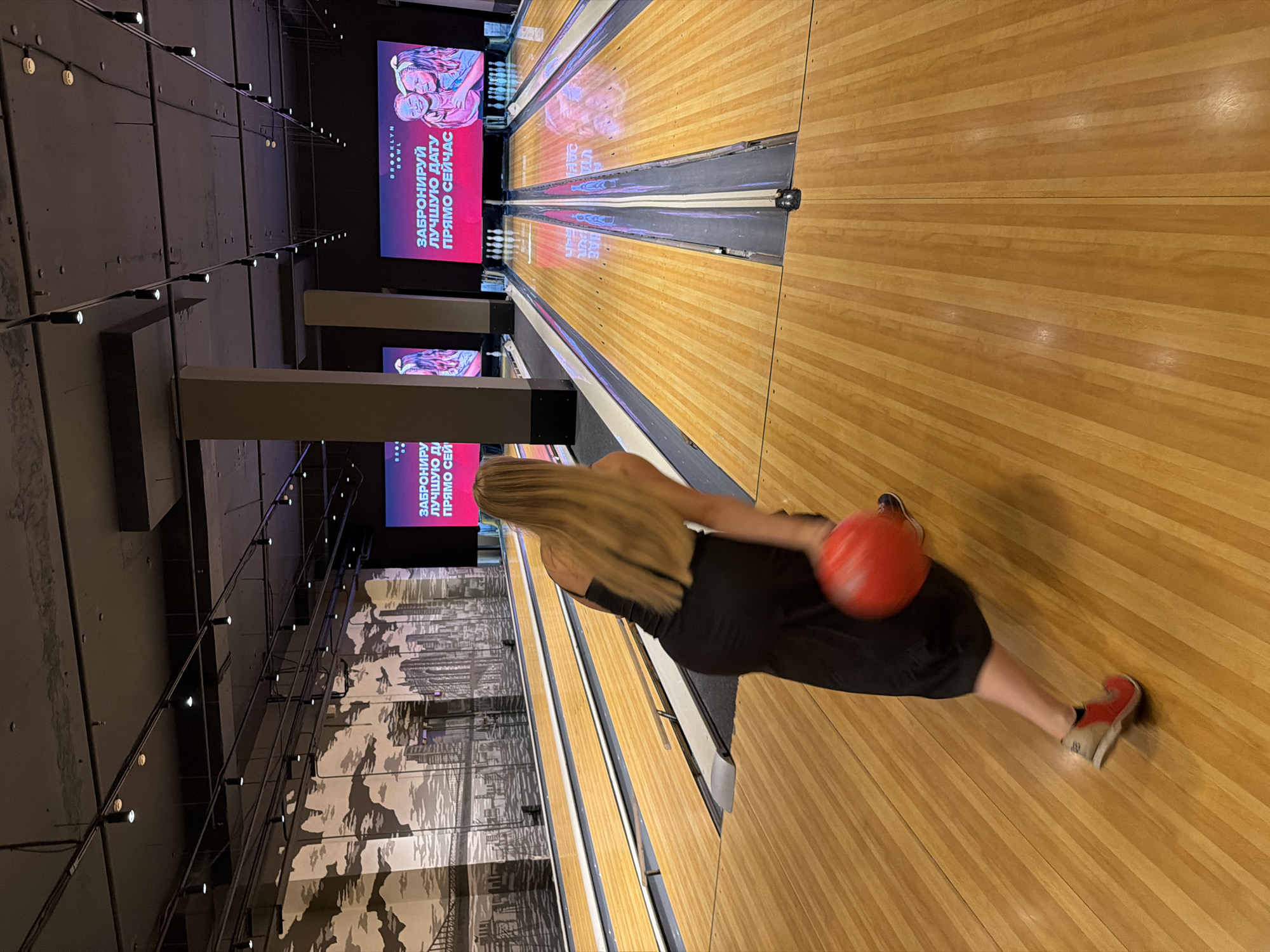 The image shows a woman bowling at "Brooklyn Bowl" in Izhevsk, Russia. She is in the foreground, wearing a black dress and red and white sneakers, and is in the process of throwing a red bowling ball down the lane. The bowling alley has a polished wooden floor and is lit by overhead lights. In the background, there are several other bowling lanes, as well as large screens displaying advertisements for the establishment. The walls are decorated with a mural depicting a city skyline. The overall atmosphere appears to be one of leisure and entertainment.