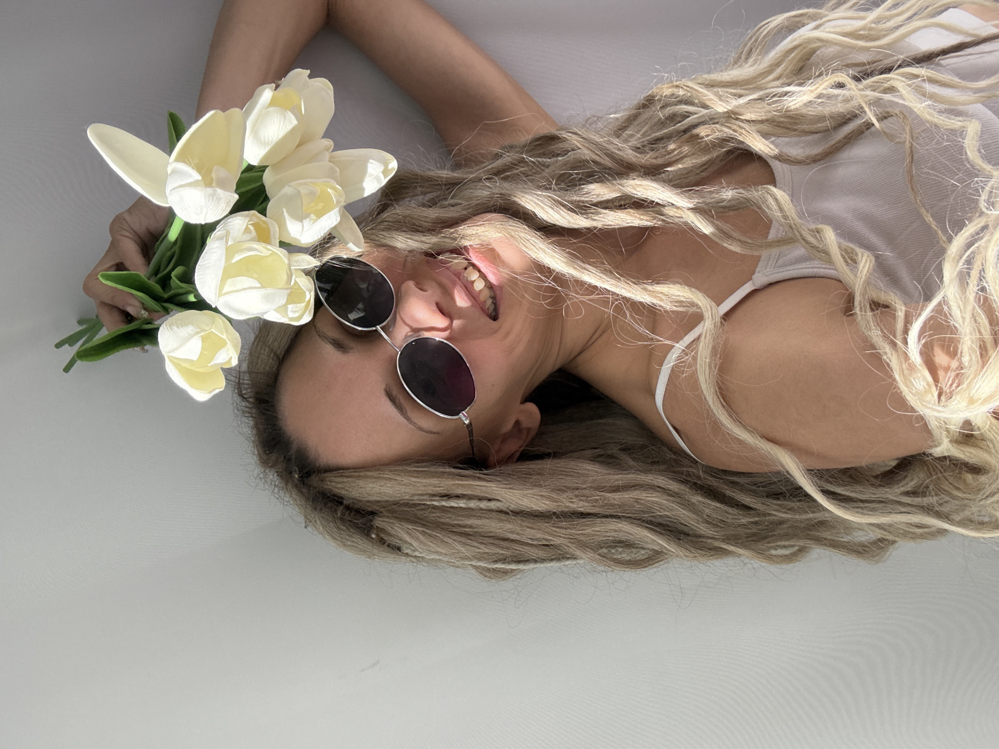 A bright, high-angle shot captures a smiling woman lying down against a plain, light-colored background, likely white or very light gray. Her body is oriented horizontally across the frame. She has long, wavy blonde hair, some strands appearing to be thin braids or dreadlocks, fanned out around her head and upper body. She is wearing dark, round-framed sunglasses and a white tank top or camisole.
Her head is tilted, and she is smiling broadly, revealing her teeth, indicating happiness and relaxation. Her left arm is raised, holding a bouquet of approximately eight to ten white or very pale yellow tulips with prominent green stems and leaves, positioned near her head. The lighting is soft and natural, casting gentle shadows that highlight the contours of her face, arms, and the flowers, suggesting a bright indoor setting or diffused outdoor light.
The composition is minimalist, focusing entirely on the woman and the flowers, conveying a sense of joy and a relaxed, summery aesthetic. There is no visible text in the image.