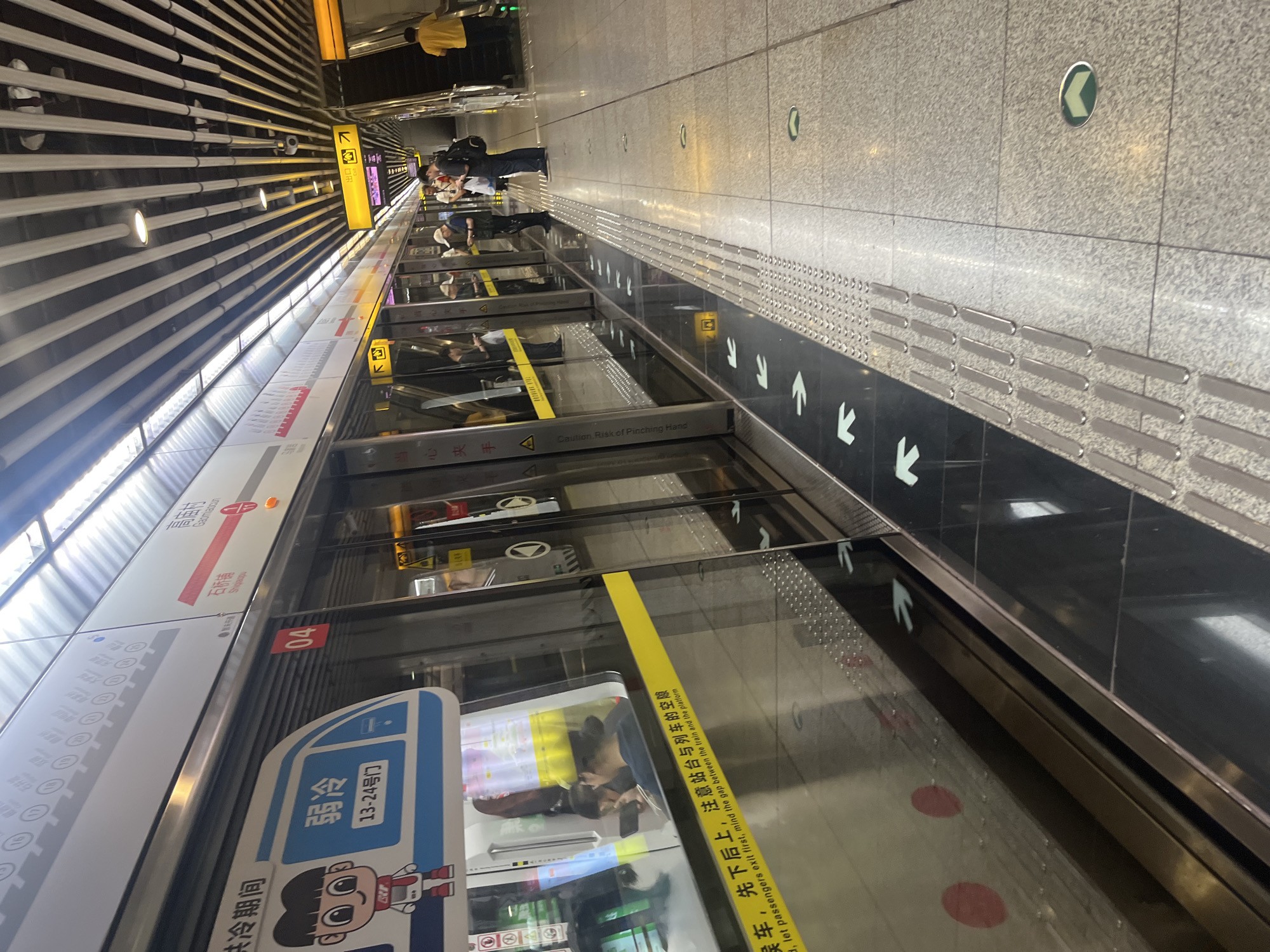 The image captures a scene at a metro station in Shapingba District, China. A train is stopped at the platform, with its doors open. On the side of the train, a sign displays "弱冷 13-24号门," indicating the door numbers for passengers. A cartoon character is also visible on the train.
Several people are present on the platform. Some are standing near the train doors, while others are further down the platform, some near an escalator. One person is wearing a yellow shirt and is near the escalator. Another person is wearing a backpack.
The platform itself is made of speckled gray tiles. There are directional arrows and tactile paving strips along the edge of the platform, indicating the safe waiting area. A yellow stripe runs along the bottom of the train doors, with Chinese characters and English text that reads, "Caution: Risk of Pinching Hand, let passengers exit first, mind the gap between the train and the platform."
The ceiling of the station features a modern design with angled metal slats and lights. Yellow signs with arrows, indicating exits, are visible overhead. The overall environment suggests a clean and functional urban transit hub.