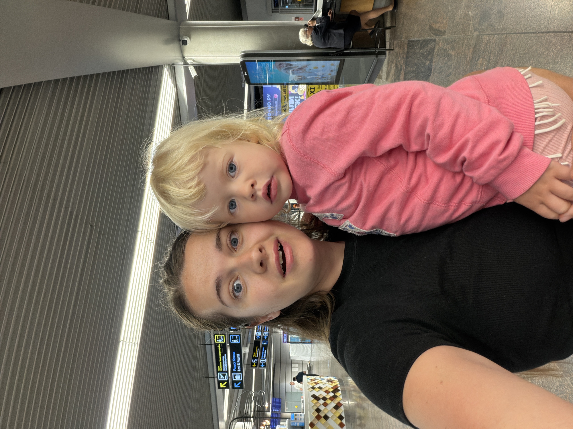 In the departure hall of Sochi International Airport, a woman with wide blue eyes and an open mouth, dressed in a black t-shirt, holds a young child with blonde hair and blue eyes wearing a pink sweatshirt and pink shorts. The child looks directly at the camera. In the background, airport signage indicates "Lost & Found" and "Domestic Departures." A woman in a dark suit sits at a counter to the right, speaking on a phone.