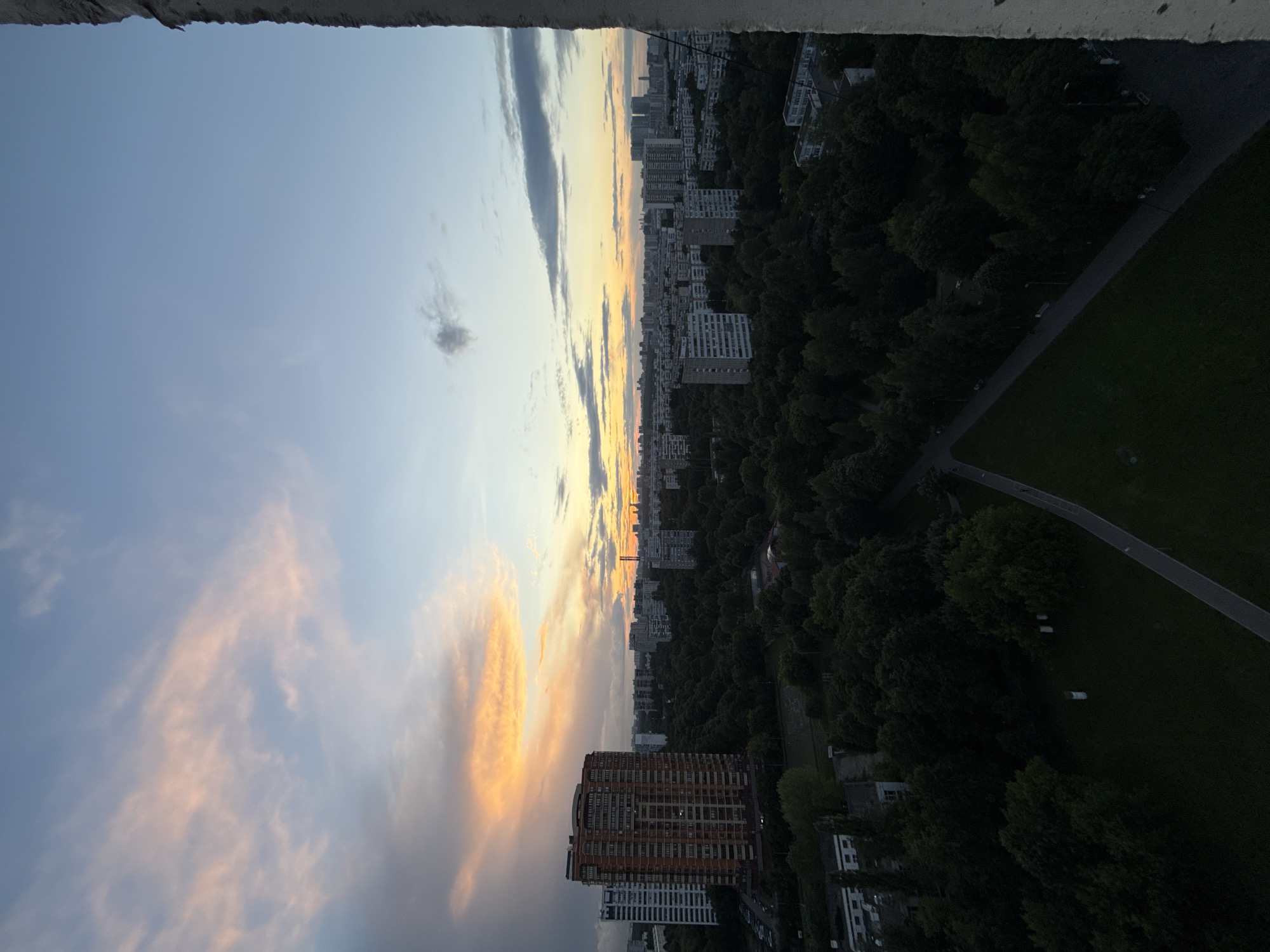 This is an aerial view of a city skyline at sunset. The image is dominated by a dramatic sky filled with streaks of orange and pink clouds against a pale blue background. In the middle ground, a dense forest of dark green trees lines a winding path. Beyond the trees, numerous tall residential buildings of varying architectural styles stretch towards the horizon. A prominent, reddish-brown apartment building stands out in the foreground. The overall scene depicts an urban environment with abundant greenery, suggesting a park or residential area with significant tree cover. The time of day is clearly sunset, as indicated by the vibrant colors in the sky and the lengthening shadows. The weather appears clear, with the sun's rays illuminating the clouds. No people are visible in the image, and there is no discernible text. The location is identified as Moscow, Russia.