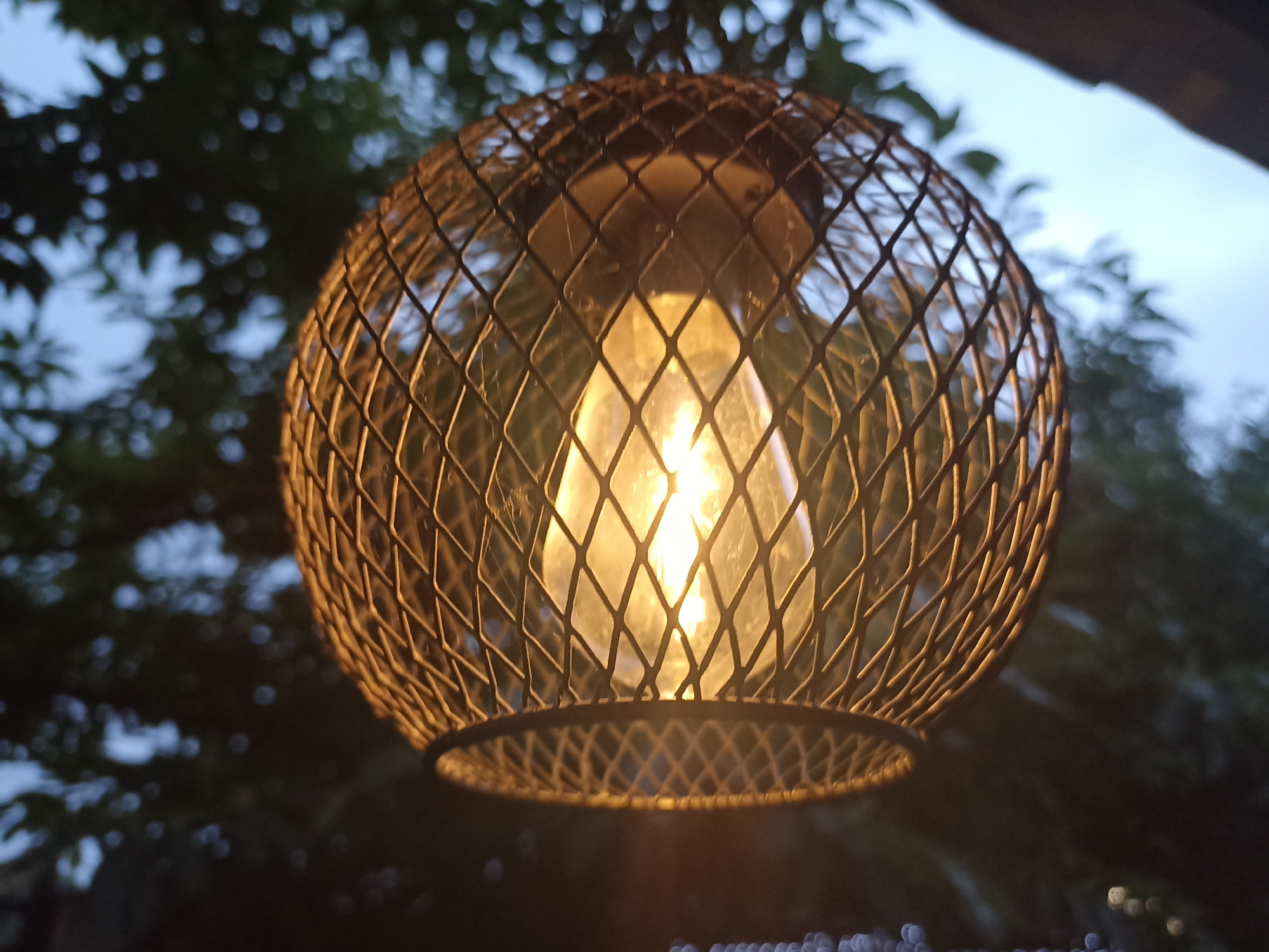 A close-up shot features a spherical wire lamp shade with a diamond-shaped mesh pattern. Inside the shade, a vintage-style incandescent light bulb glows warmly, illuminating the intricate metalwork. The background is softly blurred, showing out-of-focus dark green foliage against a pale, overcast sky. This suggests an outdoor setting, possibly a garden or patio, during daylight hours. The overall mood is cozy and atmospheric, enhanced by the soft glow of the lamp and the gentle diffusion of light through the mesh. There is no visible text in the image. The location is identified as Gusar, Azerbaijan.