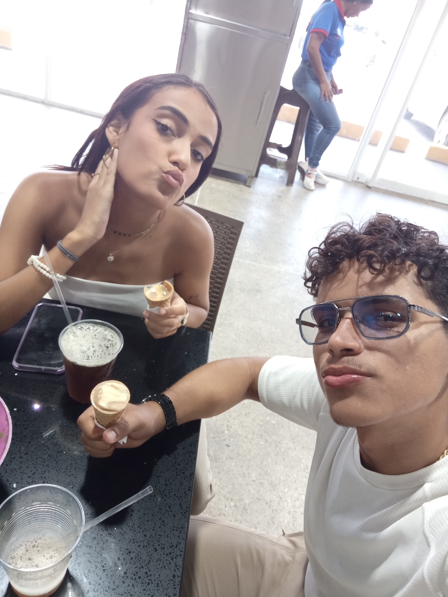 Two people are posing for a selfie in an indoor establishment. The female on the left is fair-skinned with long, dark hair, wearing a white top. She is holding an ice cream cone and has her hand near her face in a posing gesture. The male on the right is light-skinned with curly dark hair, wearing glasses and a white t-shirt. He is also holding an ice cream cone. A table is between them with beverages in cups and a cell phone. In the background, there is a woman sitting on a stool. Based on the style of the building and the people, it is likely that the location is Juan Griego, Venezuela.