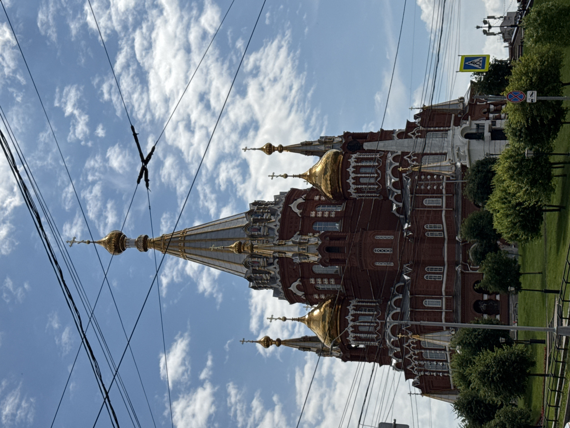 The image depicts the Saint Michael's Cathedral in Izhevsk, Russia, viewed from a low angle. The cathedral is a large, ornate brick building with multiple golden domes and spires, topped with crosses. The sky is blue with scattered white clouds. Overhead, a network of black power lines crisscrosses the frame, obscuring parts of the sky and the upper portions of the cathedral. In the foreground, there are green trees and a grassy lawn. To the right, a yellow pedestrian crossing sign and a traffic sign are visible. The overall impression is one of an urban landscape with a prominent religious structure.