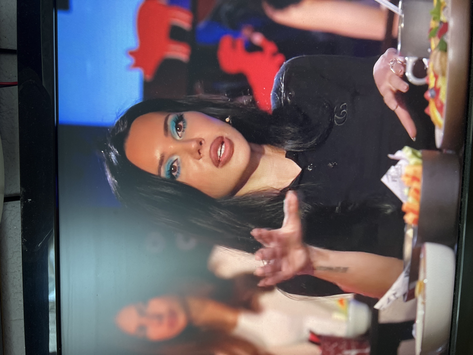 The image shows a woman with dark hair and bright blue eyeshadow. She is wearing a black shirt with a logo. She is gesturing with her left hand, which has rings on it. In the background, another woman is visible, out of focus. There are also some bowls of food on a table in front of the woman. The image is a screenshot from a television screen, indicated by the glare and the frame at the top. The provided information about the city of Kanash, Russia is not visually discernible from the image.