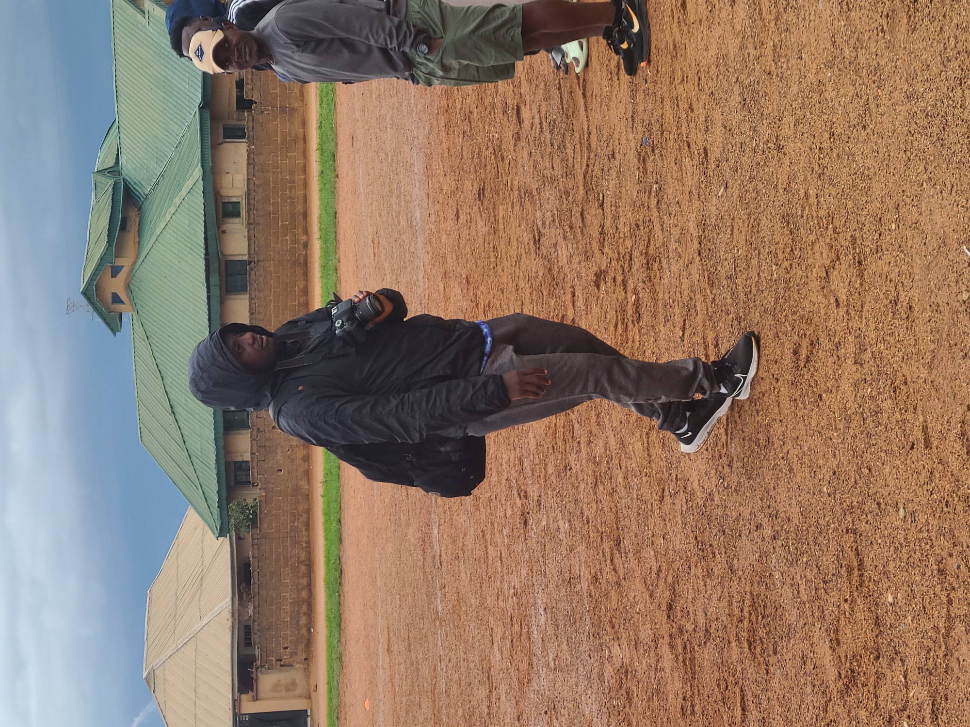 Two men are standing outdoors on a reddish-brown dirt or gravel ground. In the foreground, a man wearing a black hooded jacket, grey pants, and black sneakers with white soles holds a black DSLR camera with a lens. He also carries a black backpack. In the mid-ground, slightly to the right, another man is visible wearing a grey t-shirt, olive green shorts, black sneakers with orange accents, and a light-colored baseball cap. A third person is partially visible behind him, wearing a dark blue cap.
In the background, there are buildings with green, pitched roofs and brick walls. One building has a distinctive multi-gabled green roof. To the left of the dirt ground, a strip of vibrant green grass is visible. The sky above is light blue with some scattered clouds, indicating it is daytime, possibly overcast or partly cloudy. The overall scene suggests an outdoor event or casual gathering where photography is taking place. There is no visible text.