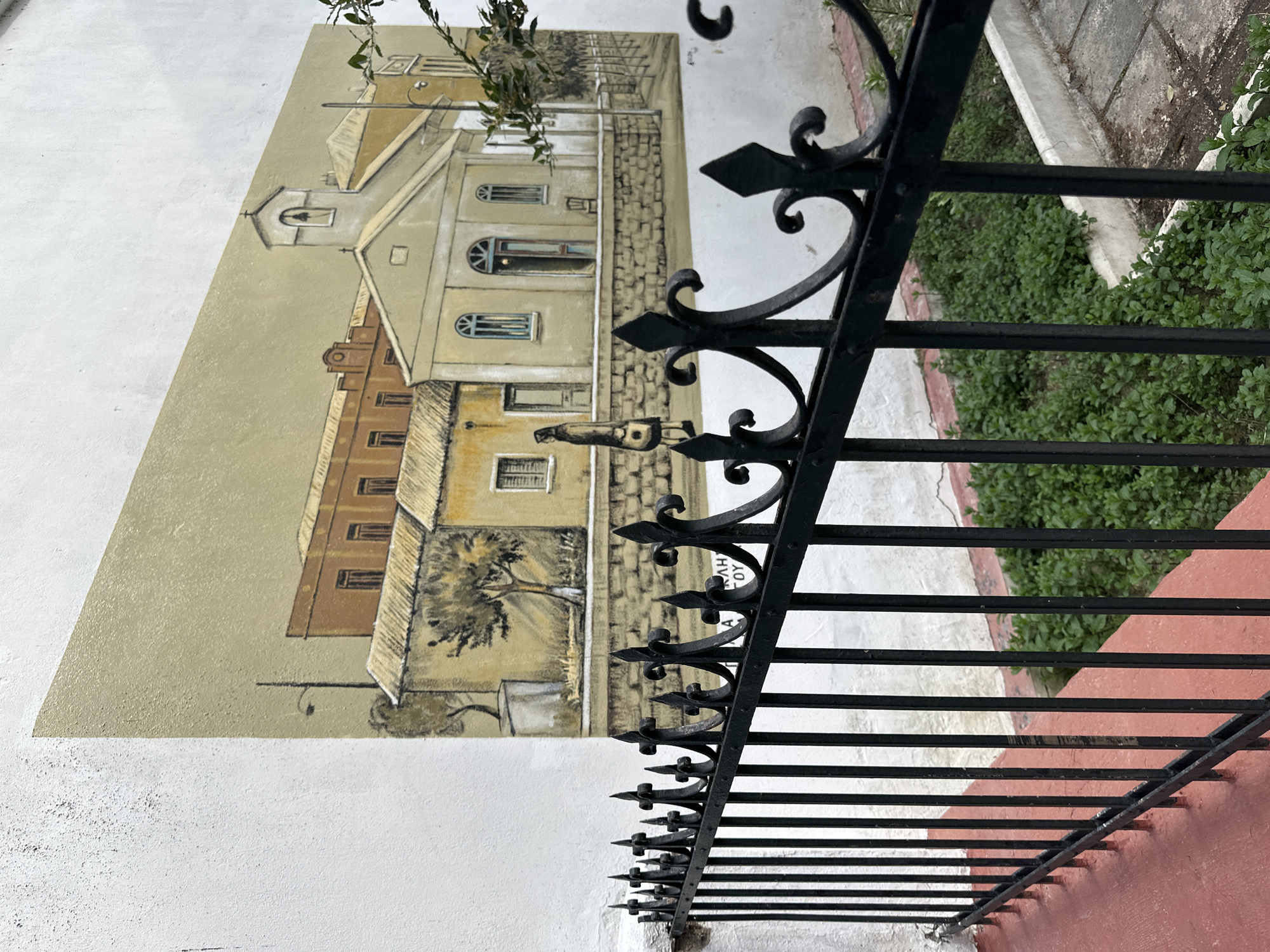 The image depicts a section of a white stucco wall in Serres, Greece, adorned with a painted mural of a cityscape in shades of beige, brown, and grey. The mural features buildings with varied architectural styles, including arched windows and a small steeple. A decorative black wrought iron fence runs parallel to the wall, partially obscuring the mural. Green ground cover is visible behind the fence, along with a glimpse of a sidewalk or path with red-painted sections. The image is taken from a ground-level perspective, looking slightly upwards and to the side.