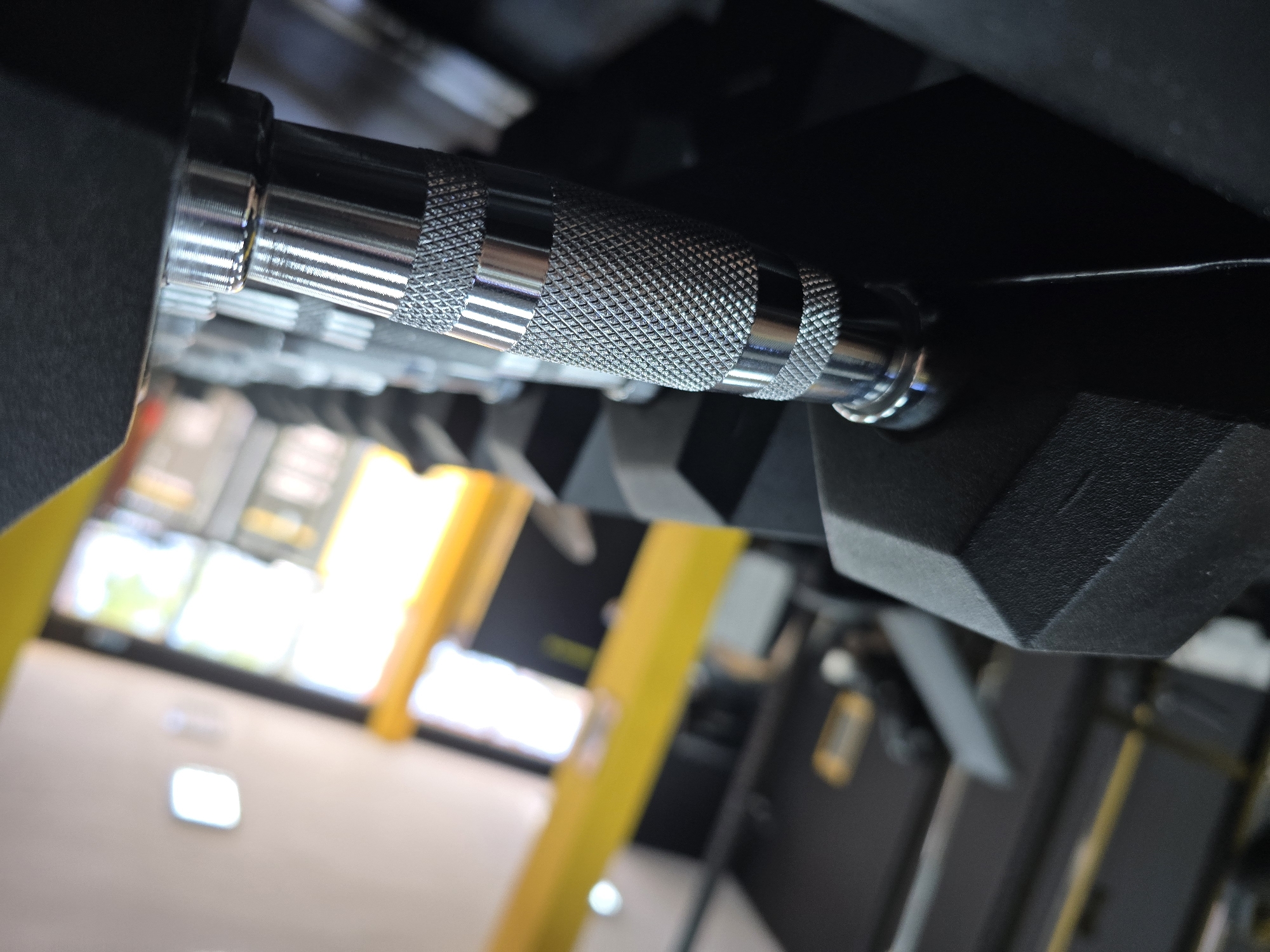 A close-up shot focuses on a set of dumbbells racked in what appears to be a gym, location unknown. The dumbbells feature knurled, chrome-colored handles. The weights themselves are black and have a multi-faceted shape. Background details are blurred, suggesting the presence of exercise equipment and a window providing natural light. The room is likely indoors.