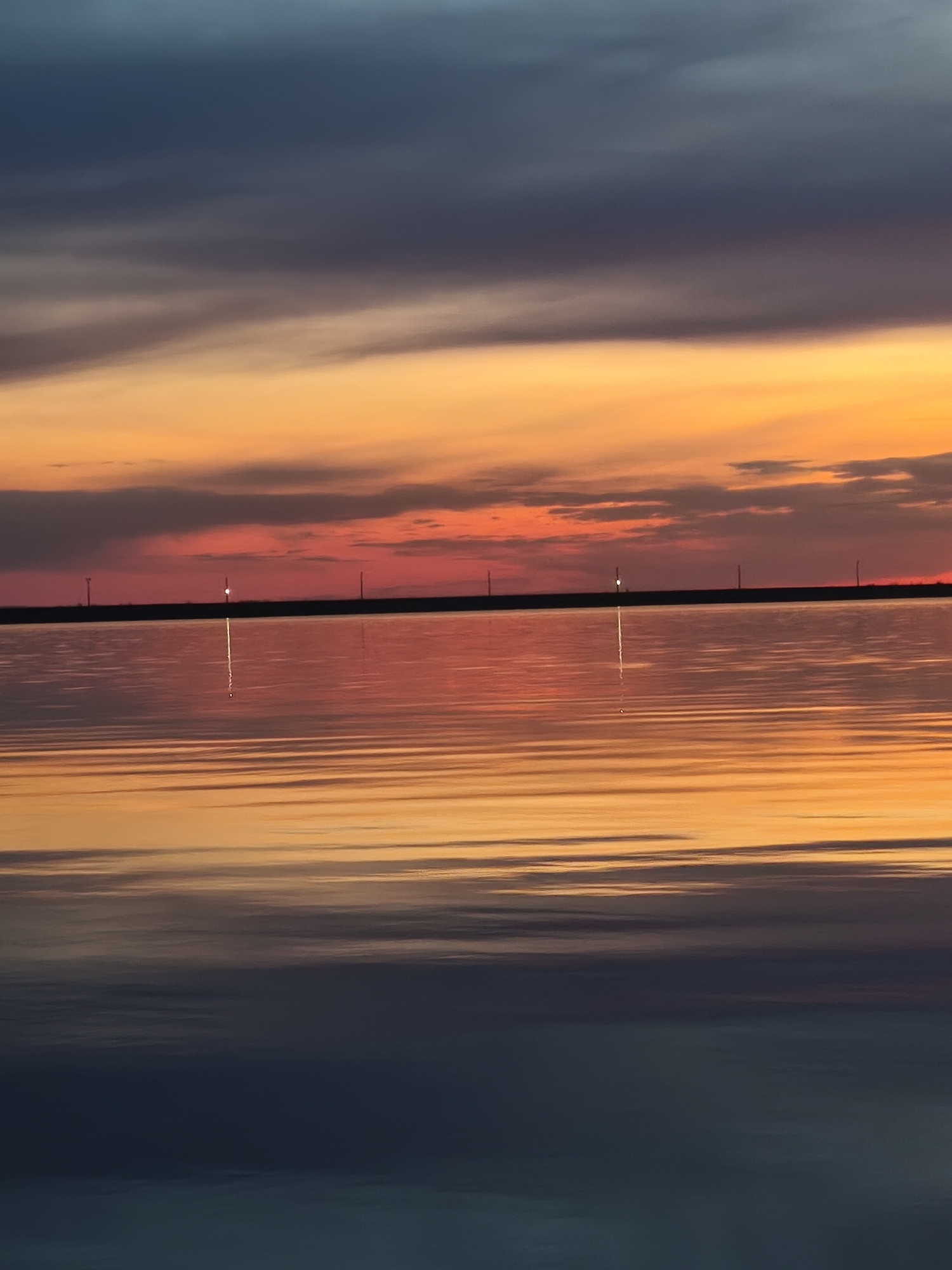 The image depicts a still body of water reflecting a vibrant sunset. The sky transitions from dark blue and gray at the top to orange and red hues near the horizon. The water mirrors these colors, creating a symmetrical effect. A dark, linear landmass is visible on the horizon, punctuated by what appear to be faint lights. The location is Chaikovsky, Russia.