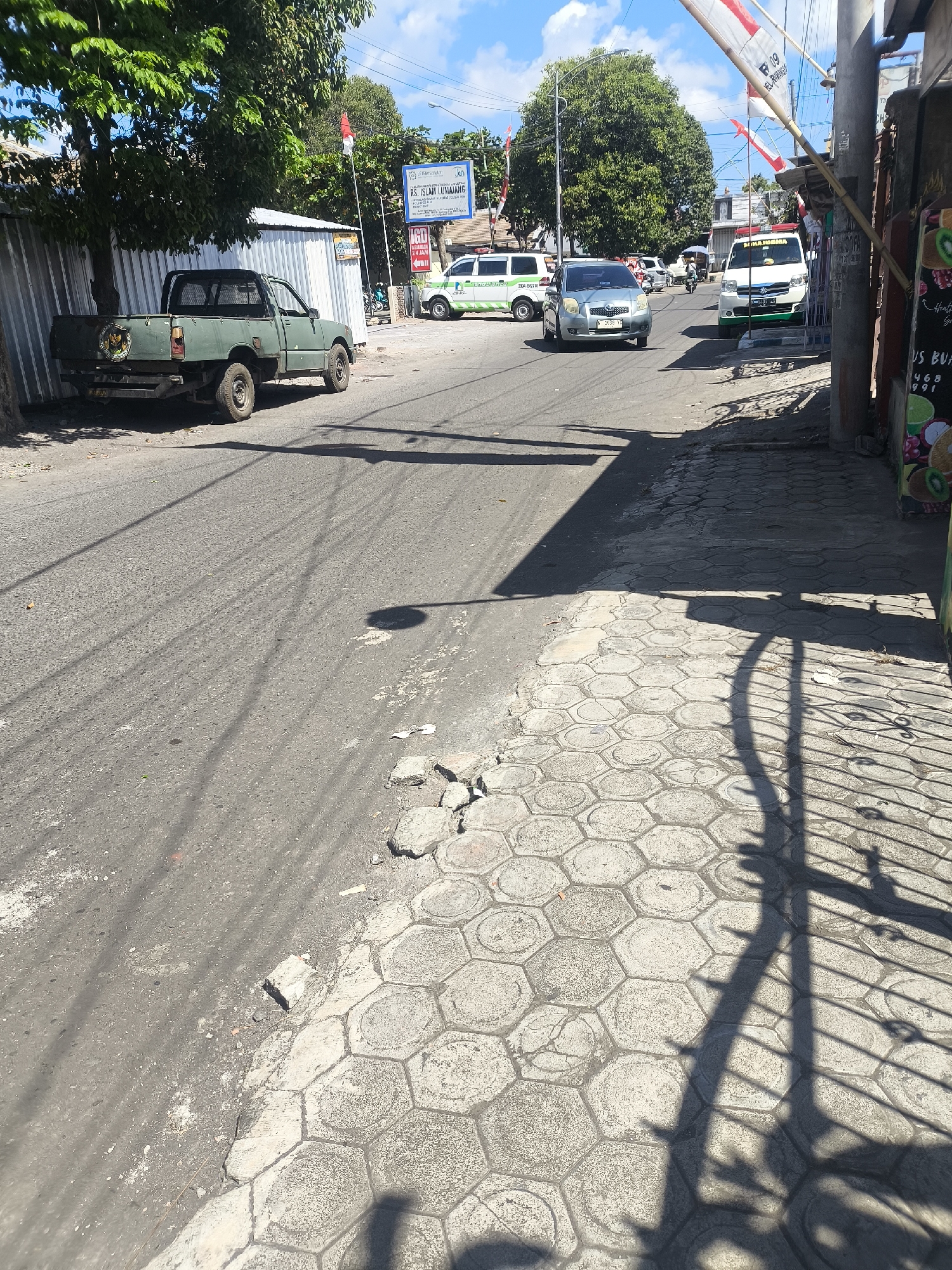The image depicts a street scene in Lumajang, Indonesia, with several vehicles visible. In the foreground, a weathered green pickup truck is parked on the left side of the road. The road itself is asphalt with a hexagonal paver sidewalk on the right. Several cars and a white van are driving on the road. In the background, there are trees, buildings, and signs, including one that appears to be for "RS ISLAM LUMAJANG". The sky is blue with some clouds, and the sun is casting distinct shadows from the vehicles and a fence on the sidewalk. The overall impression is that of a typical, slightly worn, urban street on a sunny day.