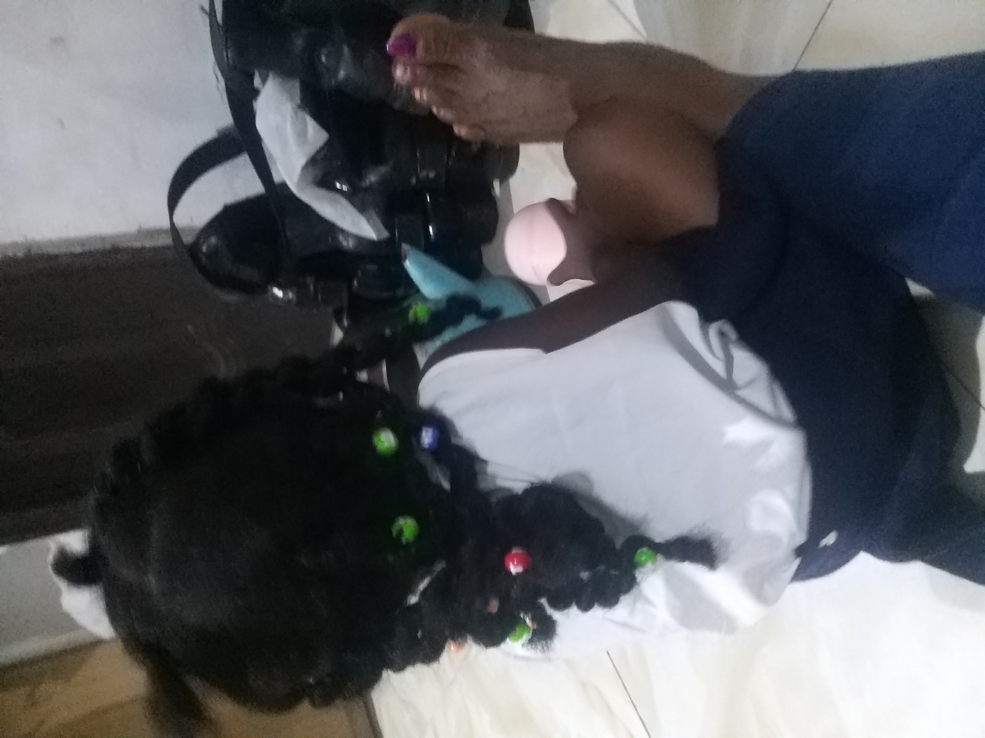 In a dimly lit interior, a person's bare foot with painted toenails rests on the floor. Adjacent to the foot, a young child with braided dark hair adorned with colorful beads is seated. The child wears a white top and dark trousers. In the background, various objects are visible, including a black and white helmet-like item and what appear to be bags. The floor is tiled. This scene is located in Goron Dutse, Nigeria.