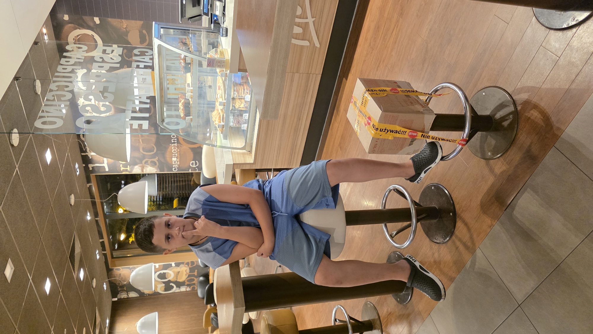 A young boy is sitting on a stool in what appears to be a McDonald's restaurant in Olempin, Poland. He is wearing a blue sleeveless shirt and blue shorts. His left hand is on his chin, and his right arm is crossed over his chest. In front of him, on another stool, is a cardboard box with yellow tape that reads "NIE UŻYWAĆ! STOP" which translates to "DO NOT USE! STOP". The background shows a typical McDonald's interior with a display case filled with pastries and drinks, seating areas, and large coffee-themed graphics on the wall.