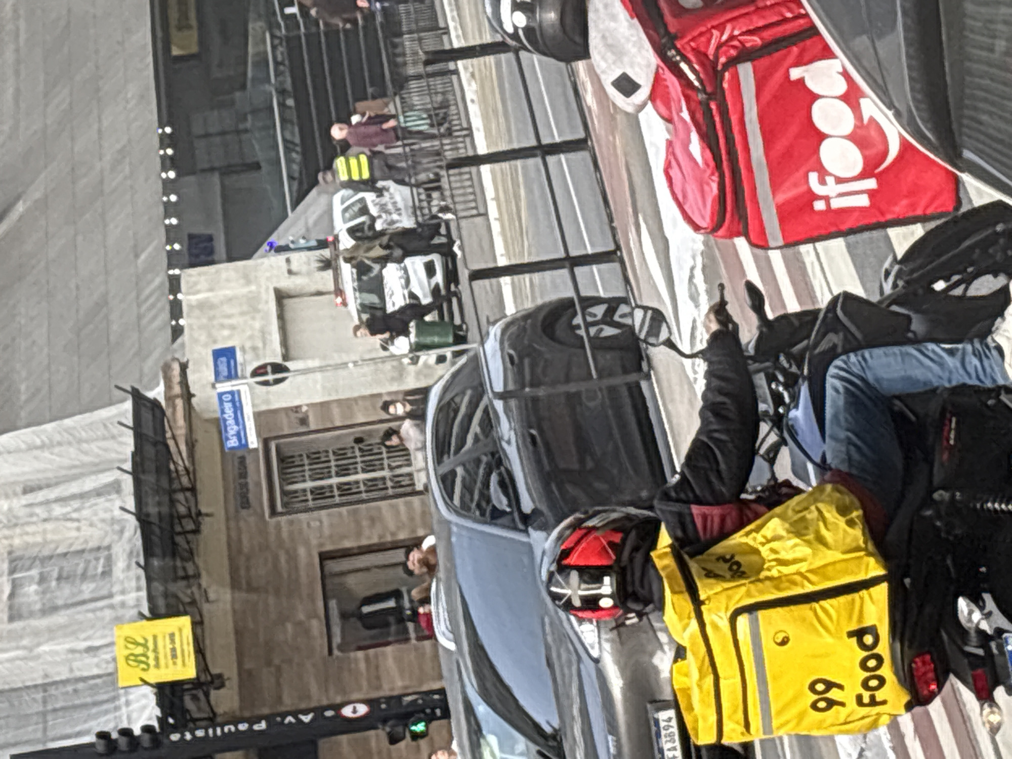 The image captures a street scene in São Paulo, Brazil, likely during daytime. In the foreground, a person on a motorcycle wears a black jacket, red and black helmet, and blue jeans, carrying a bright yellow "99 Food" delivery bag on their back. To the right, another delivery person is visible on a motorcycle with a large red "iFood" delivery bag. In the middle ground, a dark-colored SUV is parked on the street. Behind the SUV, a white police car with flashing lights is present, with several individuals standing nearby, including one in a high-visibility yellow vest. A street sign that reads "Brigadeiro" is visible. The background shows buildings and structures, with overhead wires and scaffolding. The overall environment suggests an urban setting with active traffic and commerce.