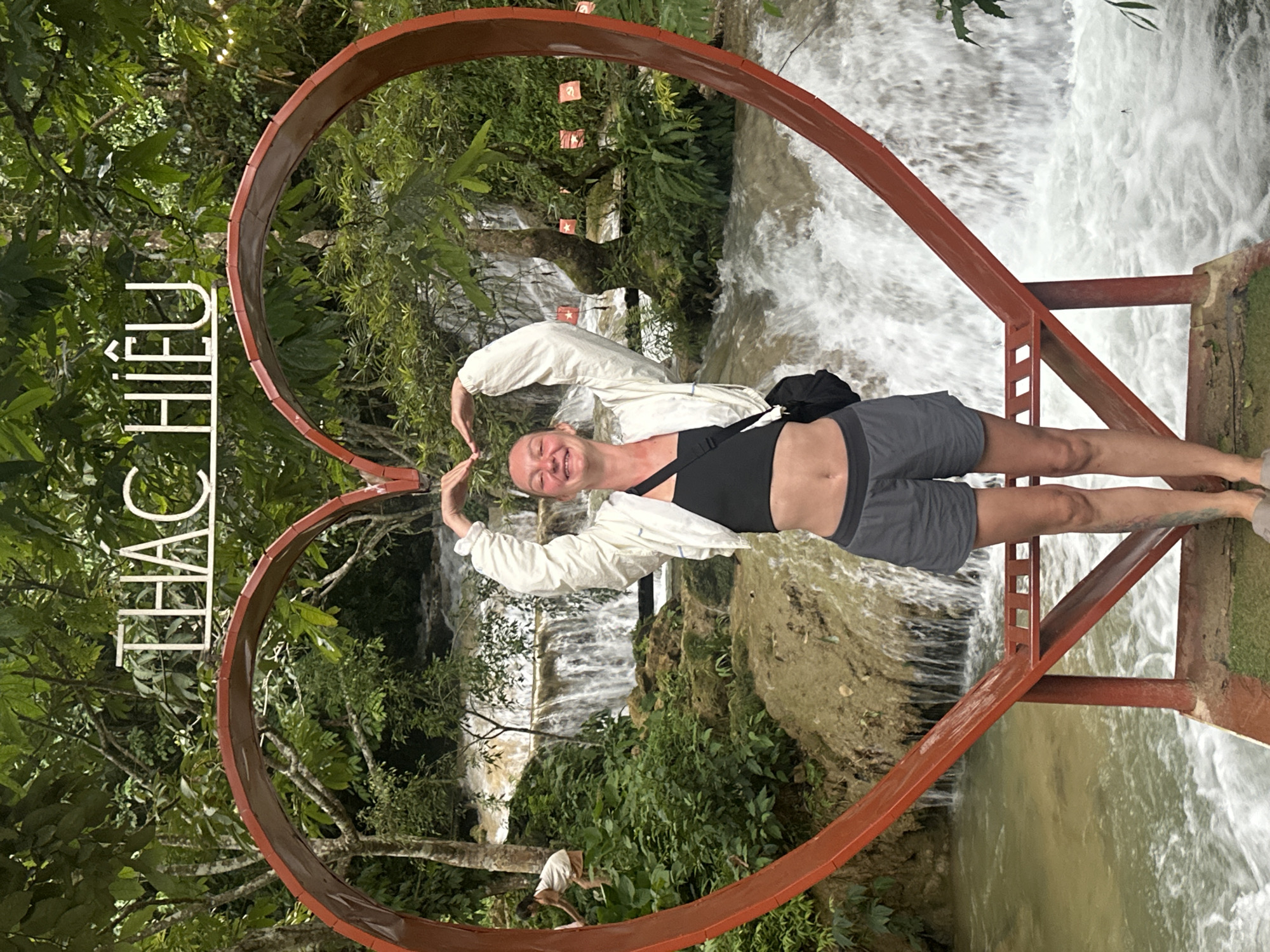In the city of Cổ Lũng Commune, Vietnam, a young woman stands in front of a waterfall, framed by a large red heart-shaped structure. She is wearing a black crop top, a white jacket, and grey shorts, with a black bag slung over her shoulder. Her hands are raised, forming a heart shape above her head. The waterfall cascades down rocks, surrounded by lush greenery. In the background, a sign with white lettering reads "THÁC HIÊU," indicating the waterfall's name. To the left, a child in a white top and brown shorts is visible near the water. Several red flags with yellow stars hang from trees in the background. The scene is set on a platform with a red railing overlooking the water.