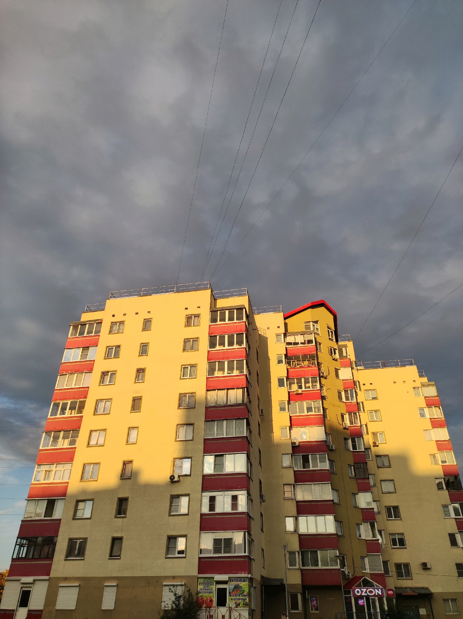 The image shows a multi-story apartment building in Chaikovsky, Russia, illuminated by the golden light of the setting sun. The building, painted in yellow and accented with red balconies and window frames, has numerous windows of various sizes. Overhead, a network of power lines stretches across a dramatic, cloudy sky. The shadow of another building is cast across the facade of the apartment building, creating a stark contrast between light and dark. The ground floor of the building features commercial establishments, including a store with "OZON" branding. There are no visible people actively engaged in any activity in the frame. The scene captures a typical urban landscape with residential and commercial elements under a dynamic sky.