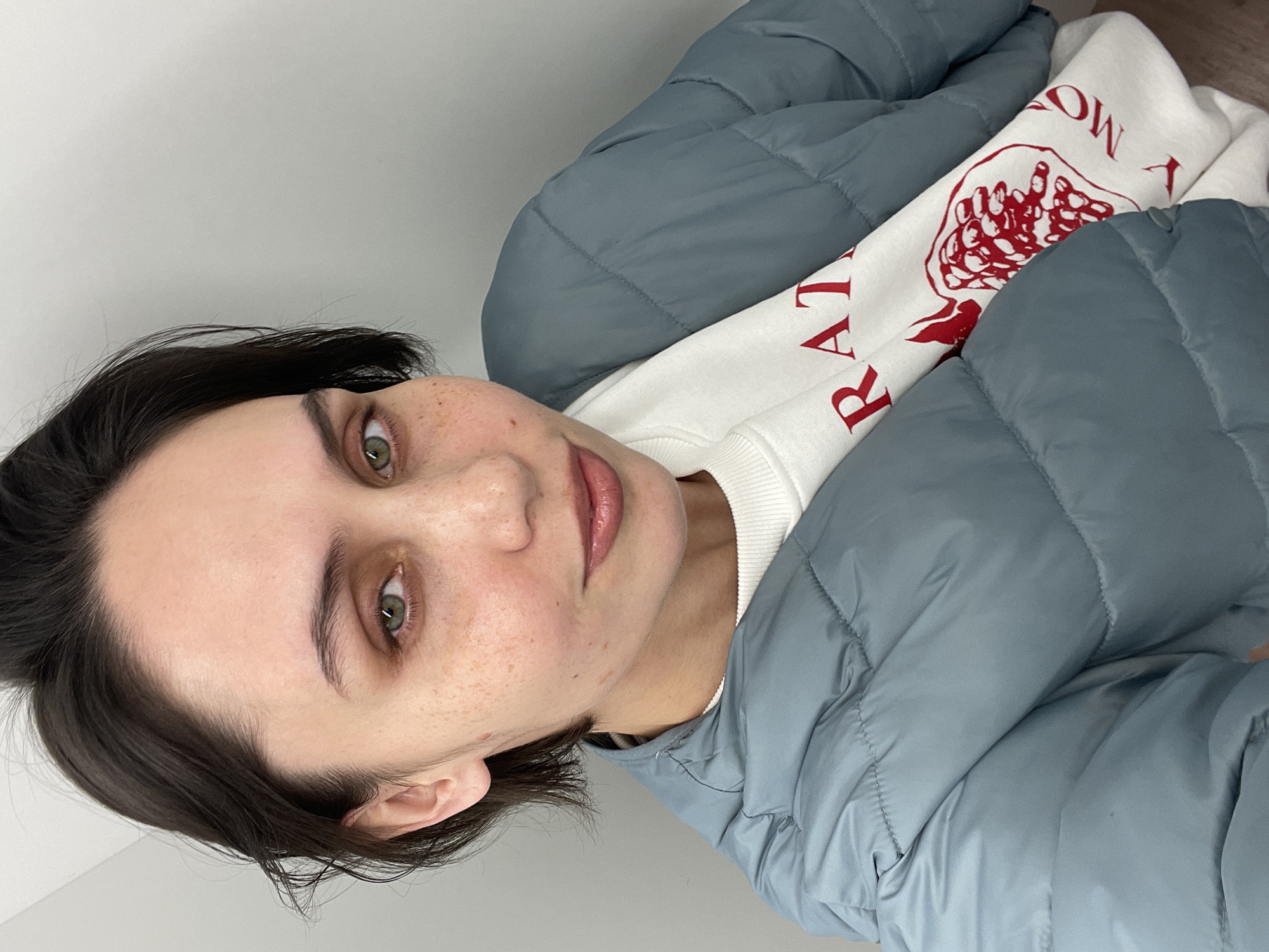 A woman with short dark hair and green eyes is wearing a light blue quilted jacket over a white sweatshirt with red lettering and a pomegranate design. She is looking at the camera. The location is not discernible as Izhevsk, Russia, or any other specific city. The background appears to be a plain white wall.