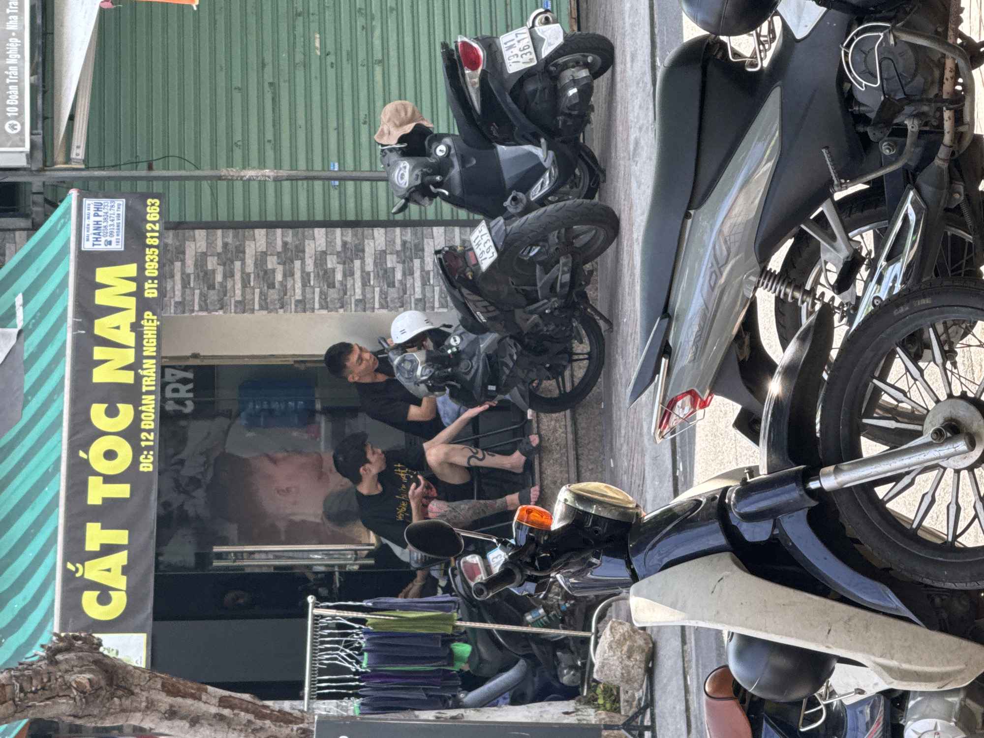 The image depicts a street scene in Bac Nha Trang Ward, Vietnam, featuring a barbershop with the sign "CẮT TÓC NAM" (Men's Haircut) prominently displayed. Two men are seated on the steps outside the shop, one appearing to be a customer with visible tattoos on his leg, and the other, possibly an employee. Several motorcycles are parked in front of the establishment, with one person sitting on a motorcycle. In the foreground, another motorcycle is partially visible. The barbershop has a modern facade with stone tiling and large glass windows. The background shows a closed green metal shutter, typical of storefronts in the area. The overall environment suggests a busy commercial street.